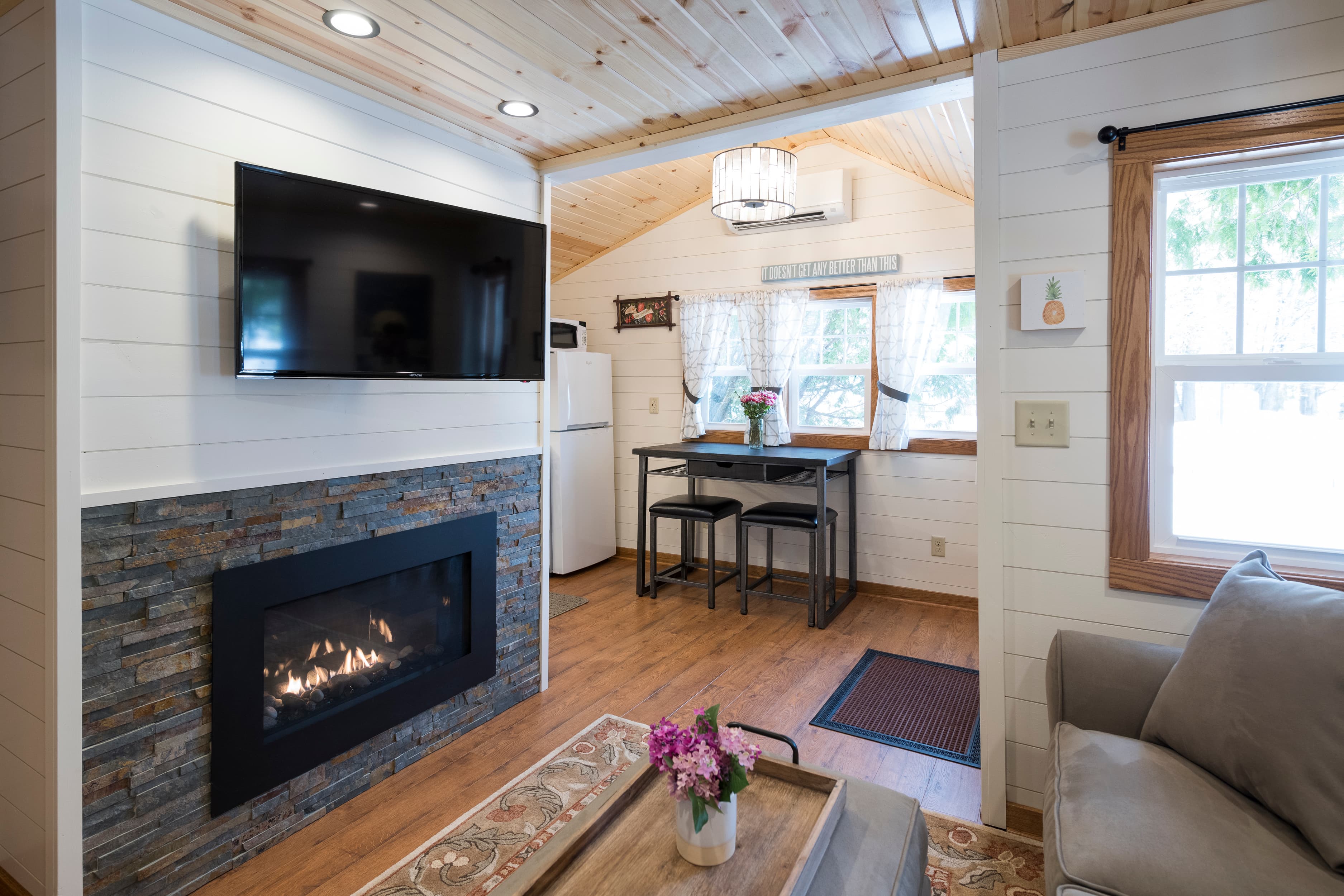 A modern living area with a fireplace and mounted TV, a dining nook, and a kitchen visible, all beneath a light wood-paneled ceiling.