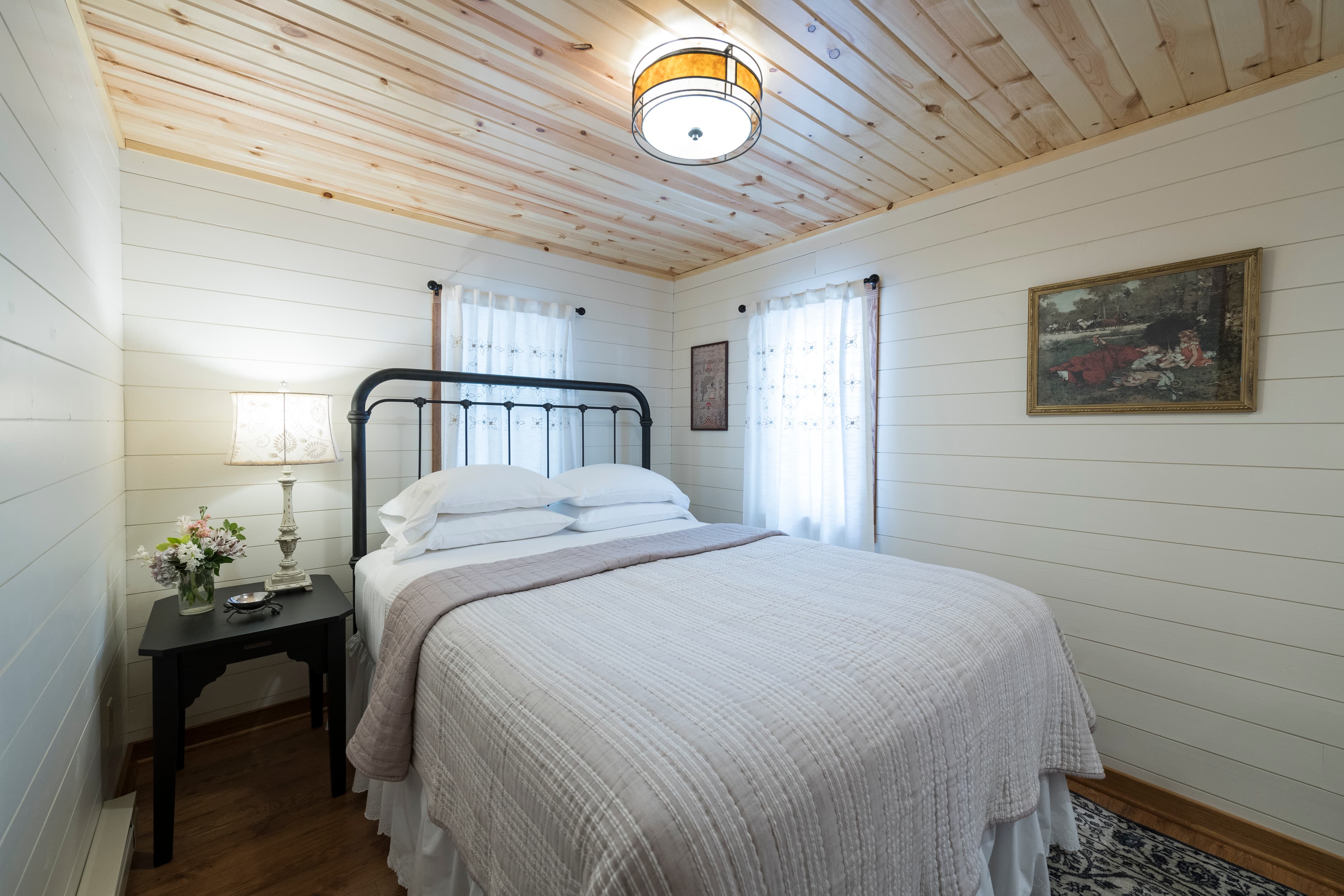 A cozy bedroom featuring white shiplap walls, a light wood ceiling, a metal frame bed with white bedding, and a small nightstand.