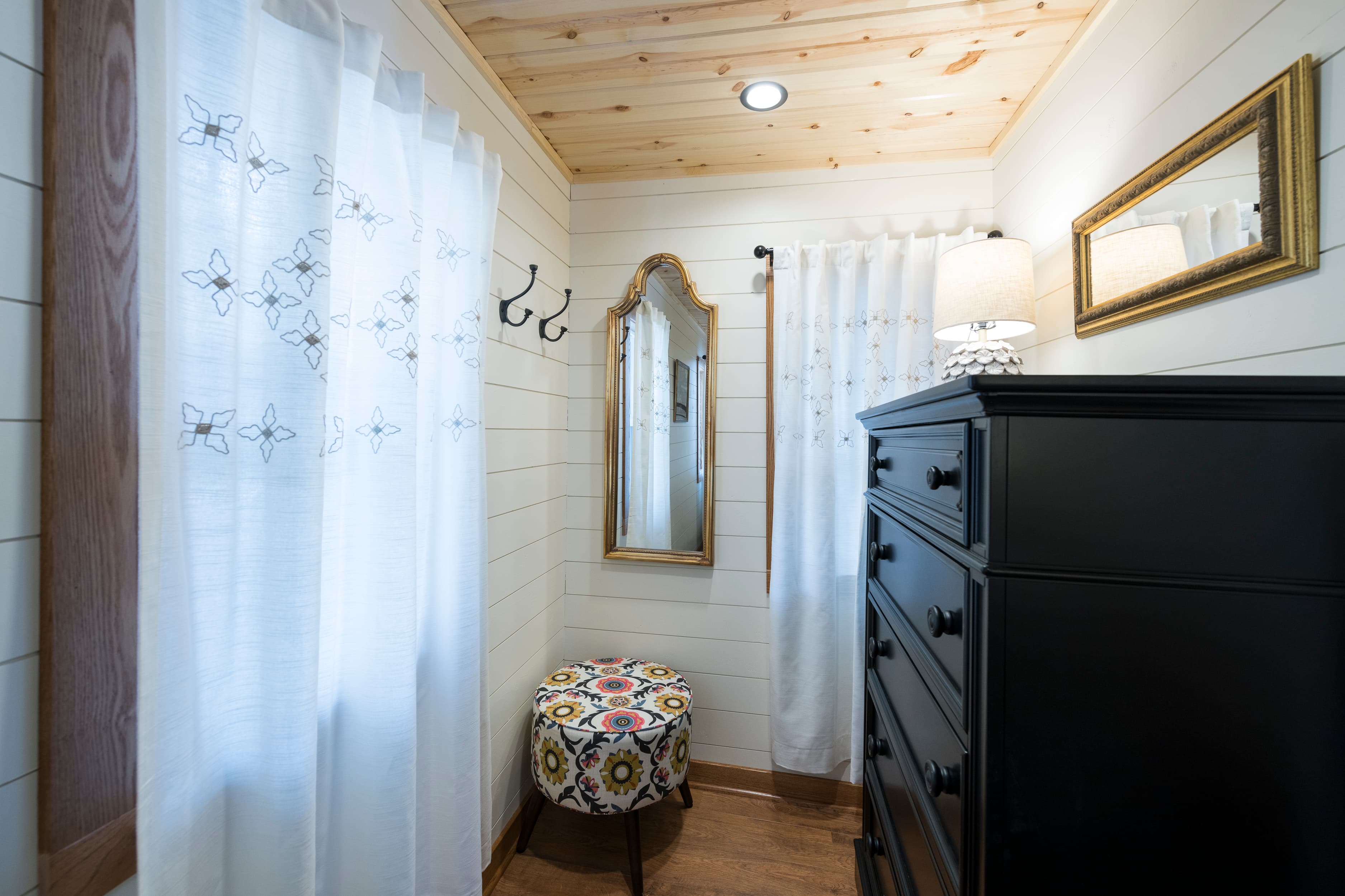 A small dressing area with white shiplap walls, a wooden ceiling, a black dresser, and a decorative stool with a colorful pattern.