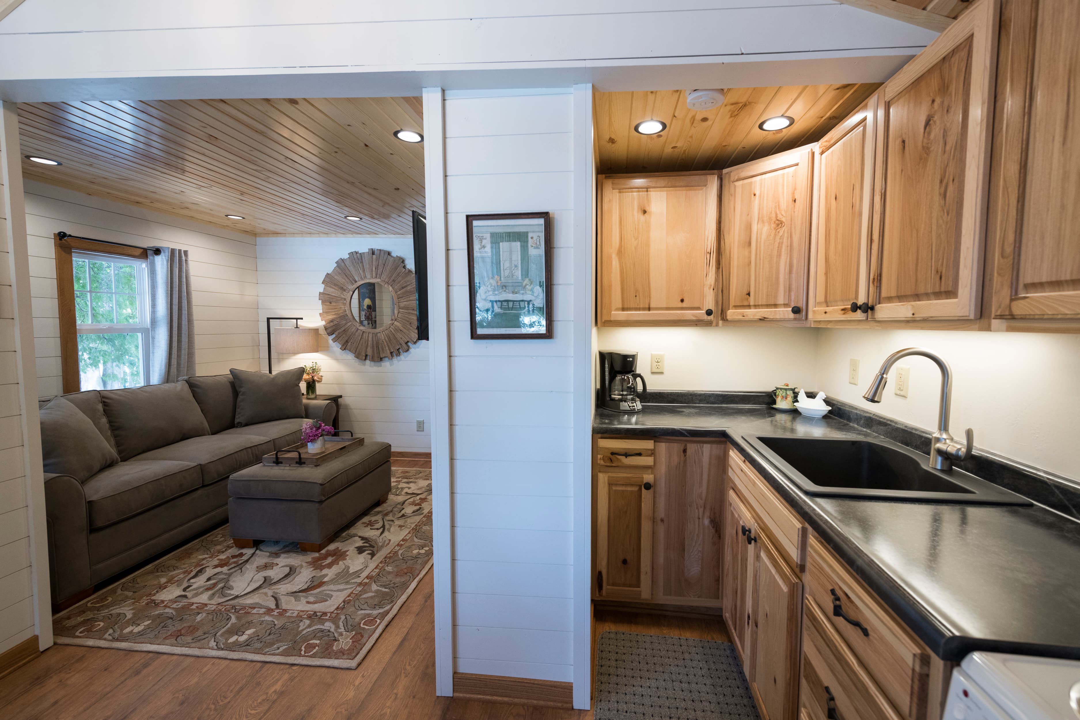 A well-lit kitchen with wooden cabinets and a dark countertop, partially open to a cozy living room with a sofa and rug.