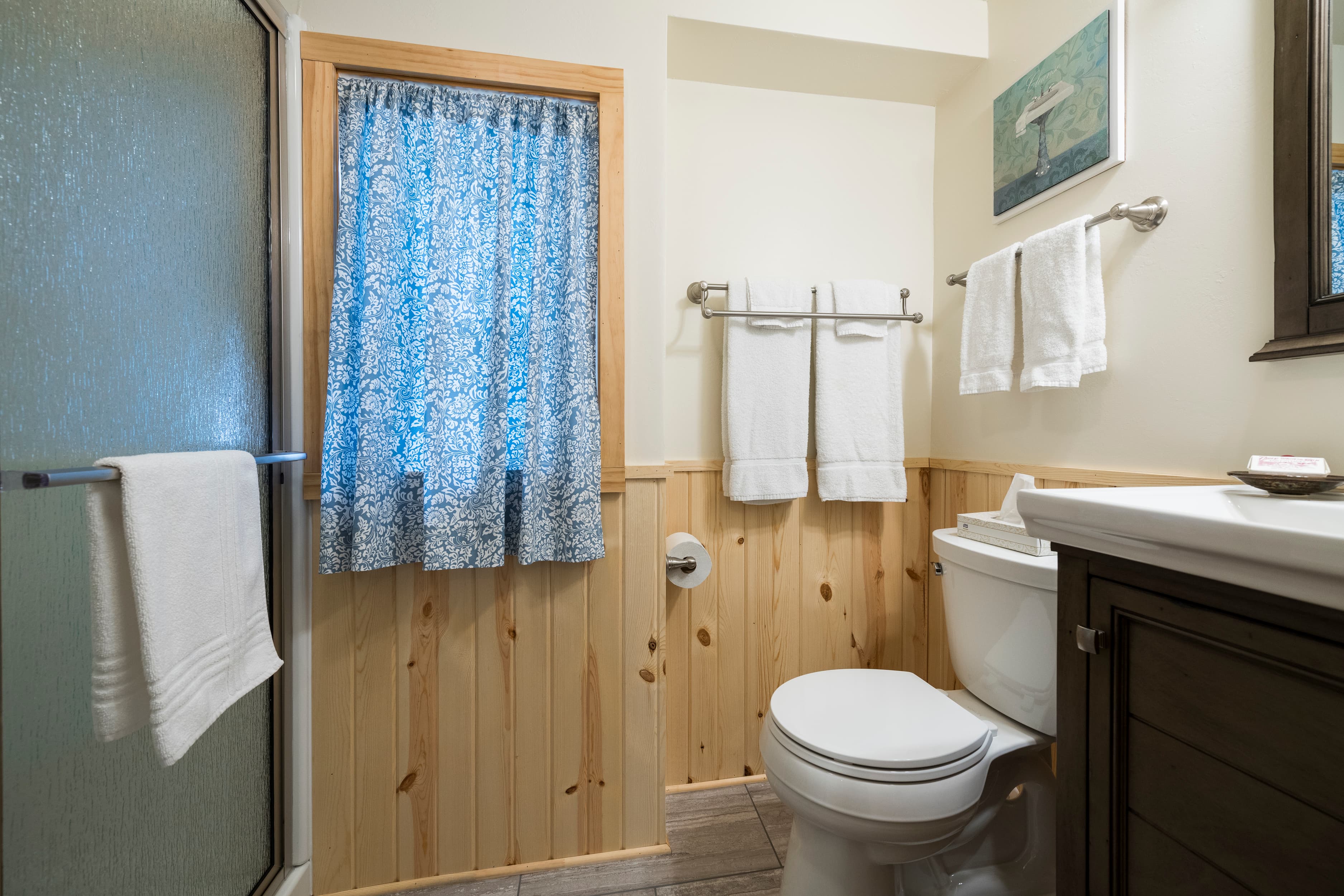 A cozy bathroom featuring light wood paneling, a white toilet, a shower with a clear glass door, and a window with a blue patterned curtain.