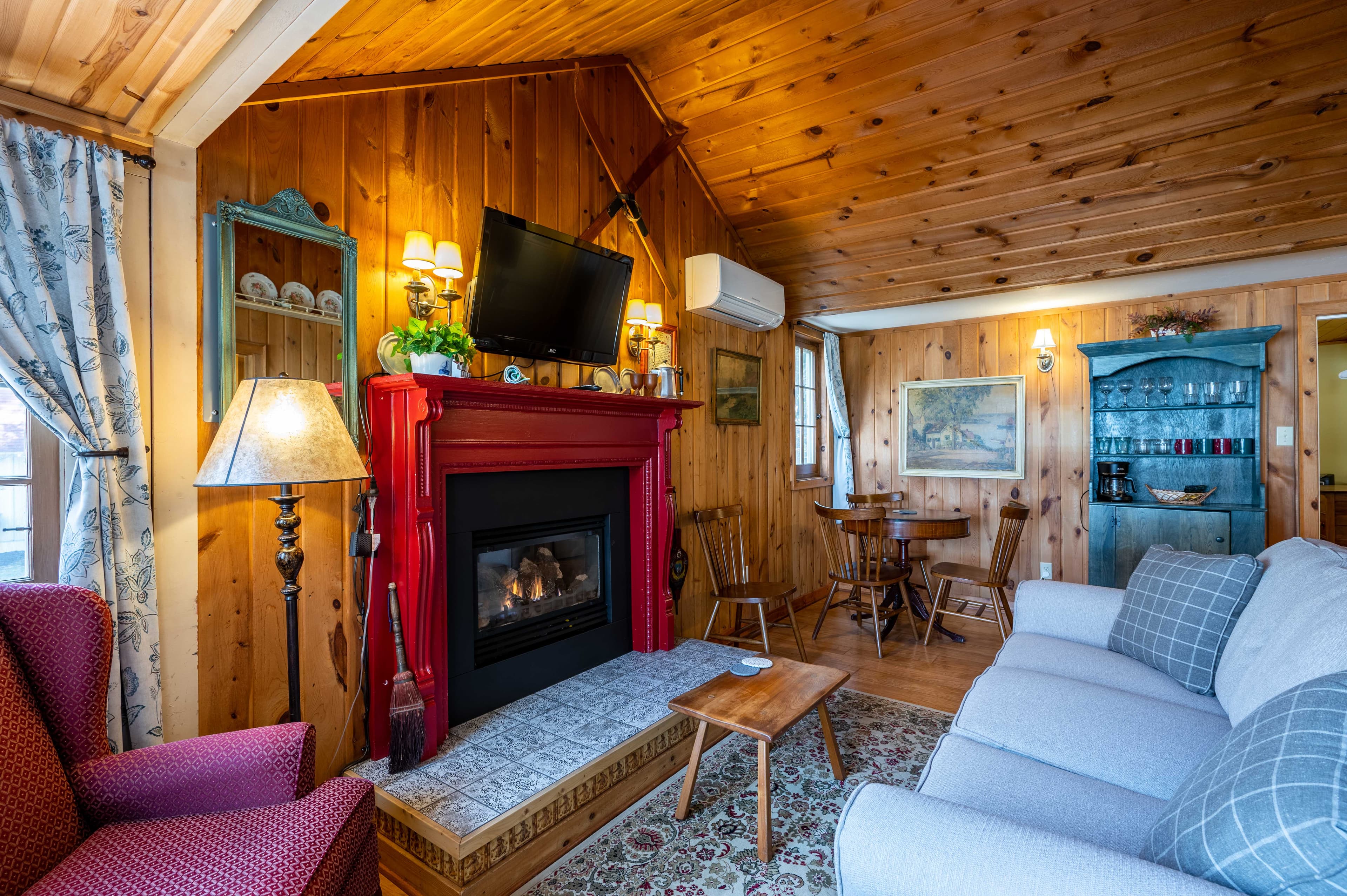 A rustic living room with wood-paneled walls and ceiling, a red fireplace, a white sofa, and a cozy armchair.