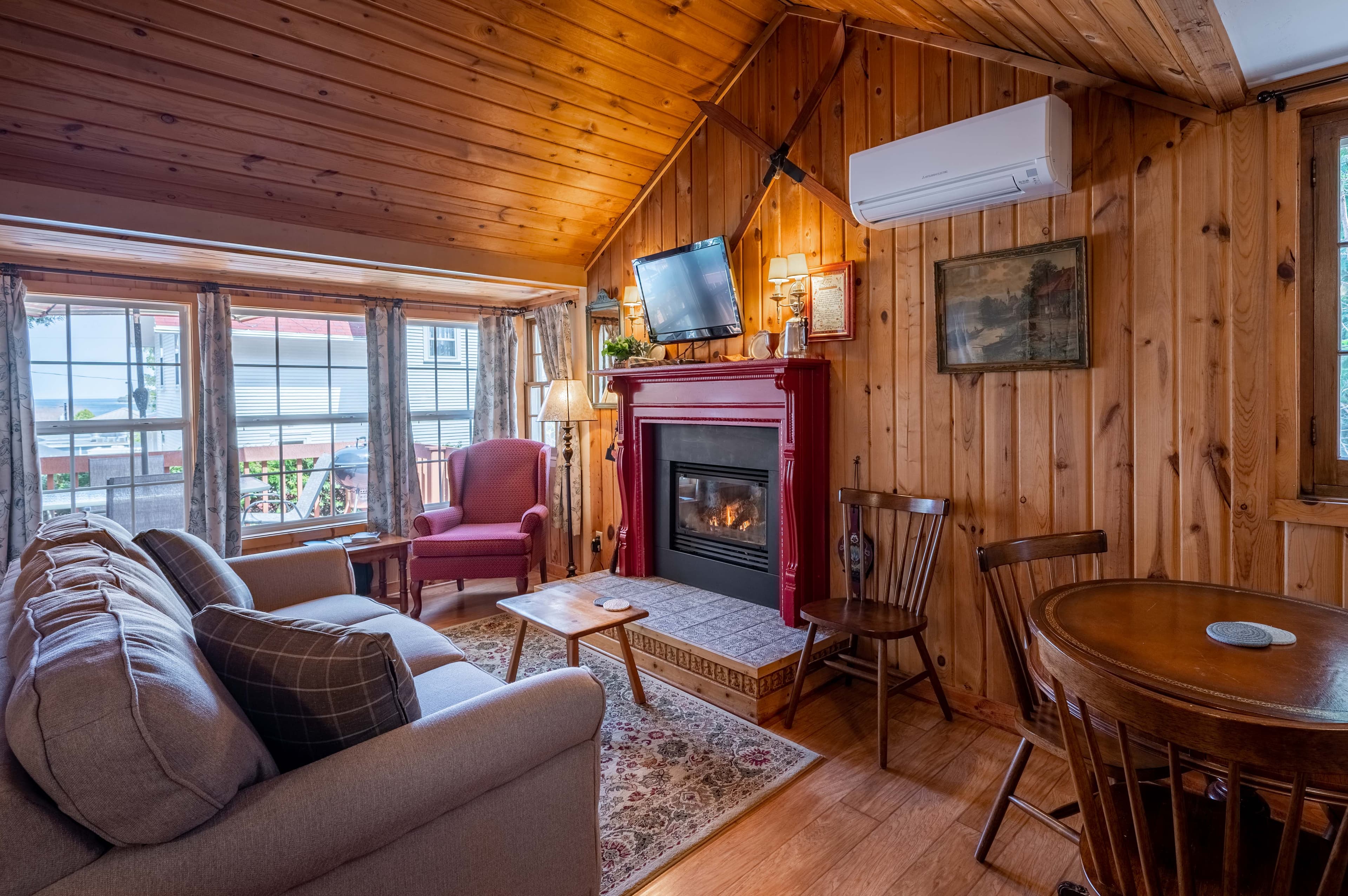 A cozy, rustic living room with wood-paneled walls and ceiling, a red fireplace, a comfortable sofa, and a dining table.