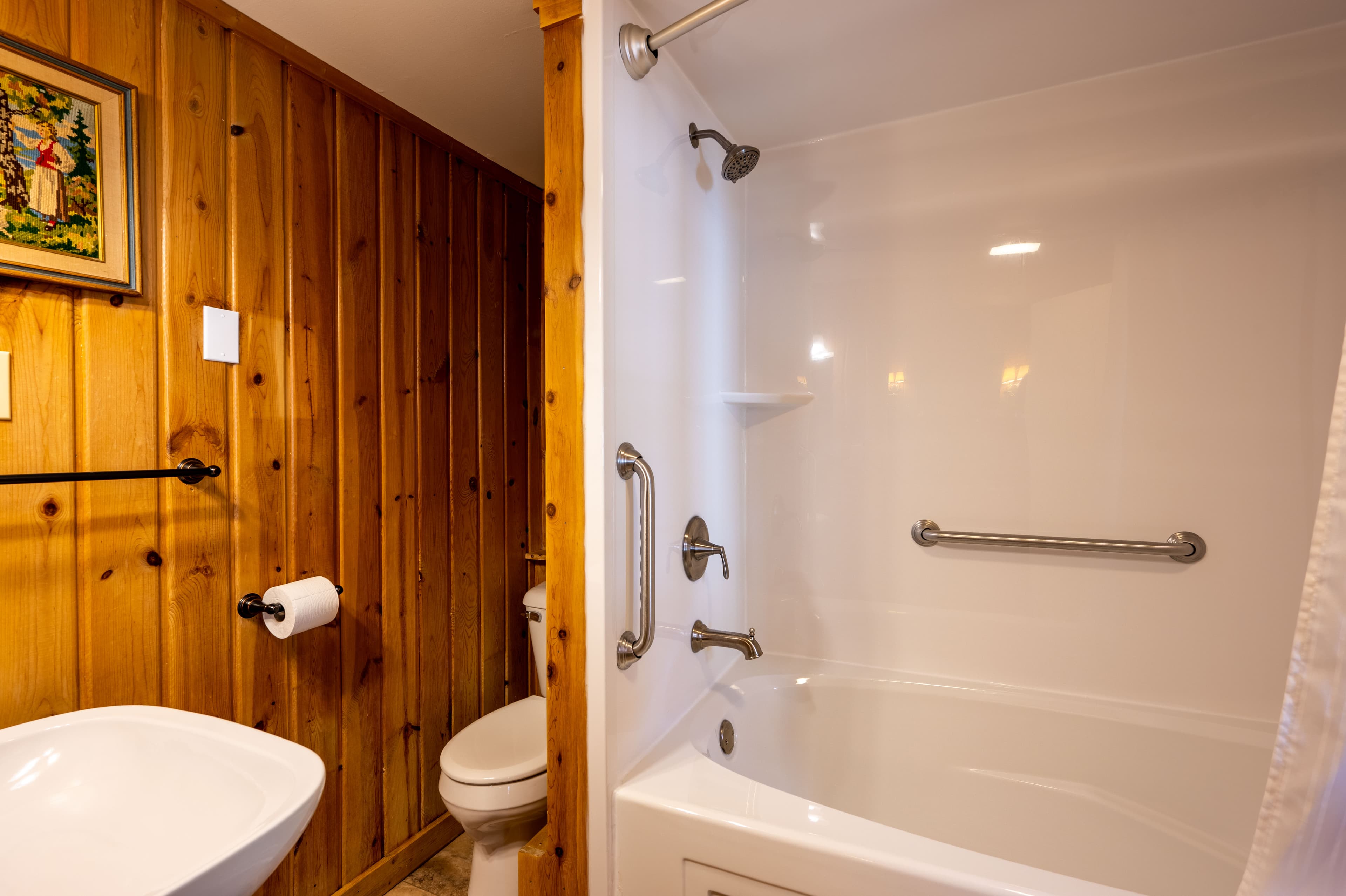 A functional bathroom featuring warm wood paneling, a white sink, a toilet, and a bathtub with a shower and grab bars.