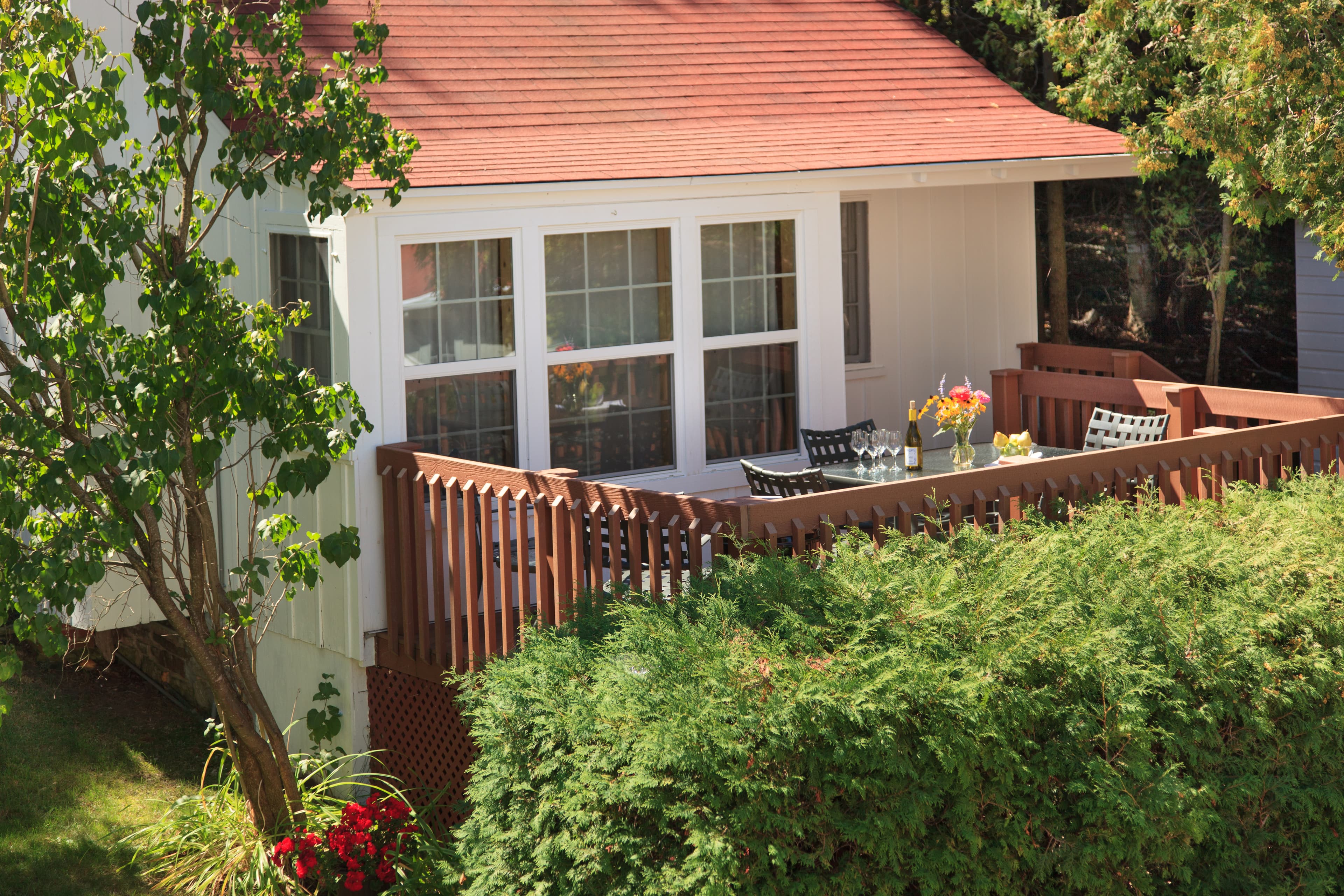 A quaint white cottage with a red roof and a spacious wooden deck furnished with outdoor seating, surrounded by vibrant green foliage.
