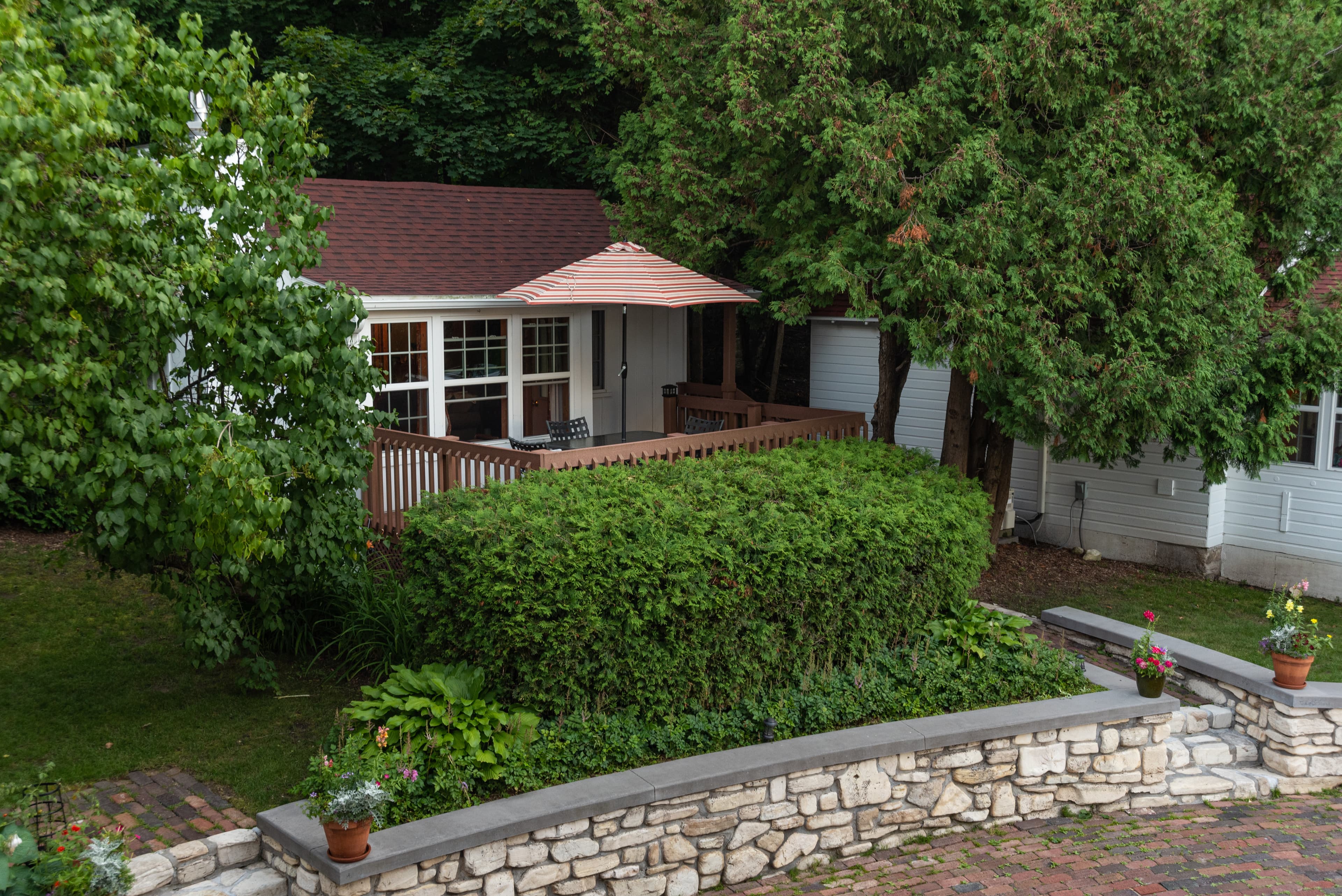 A cozy cottage with a red roof and a wooden deck featuring an umbrella, nestled amidst lush green trees and a stone wall.