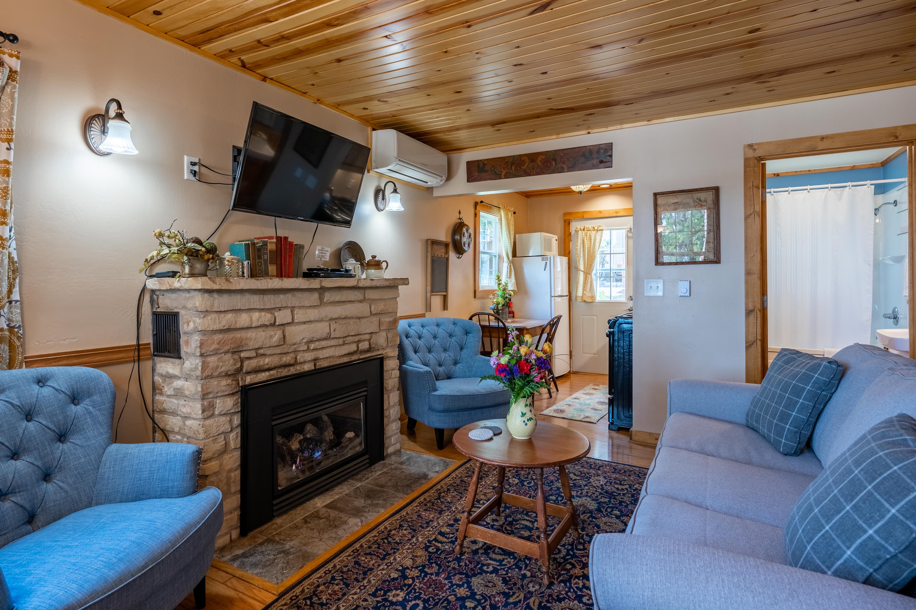 A welcoming living room with a stone fireplace, a wall-mounted TV, a blue armchair and sofa, and warm wood-paneled ceilings.