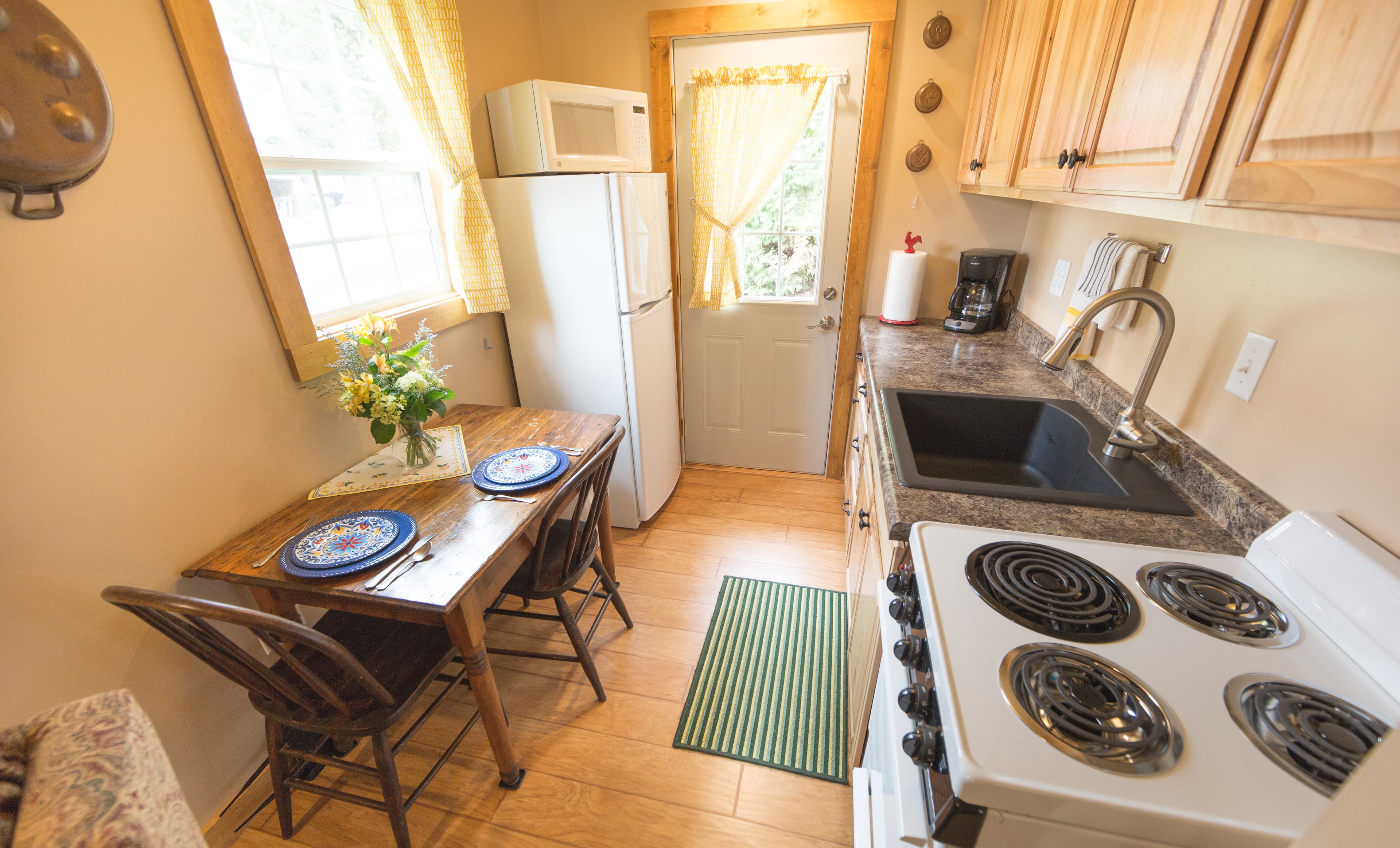 A small, functional kitchen with light wood cabinets, a white stove, a dining table for two, and a bright yellow curtain on the window.