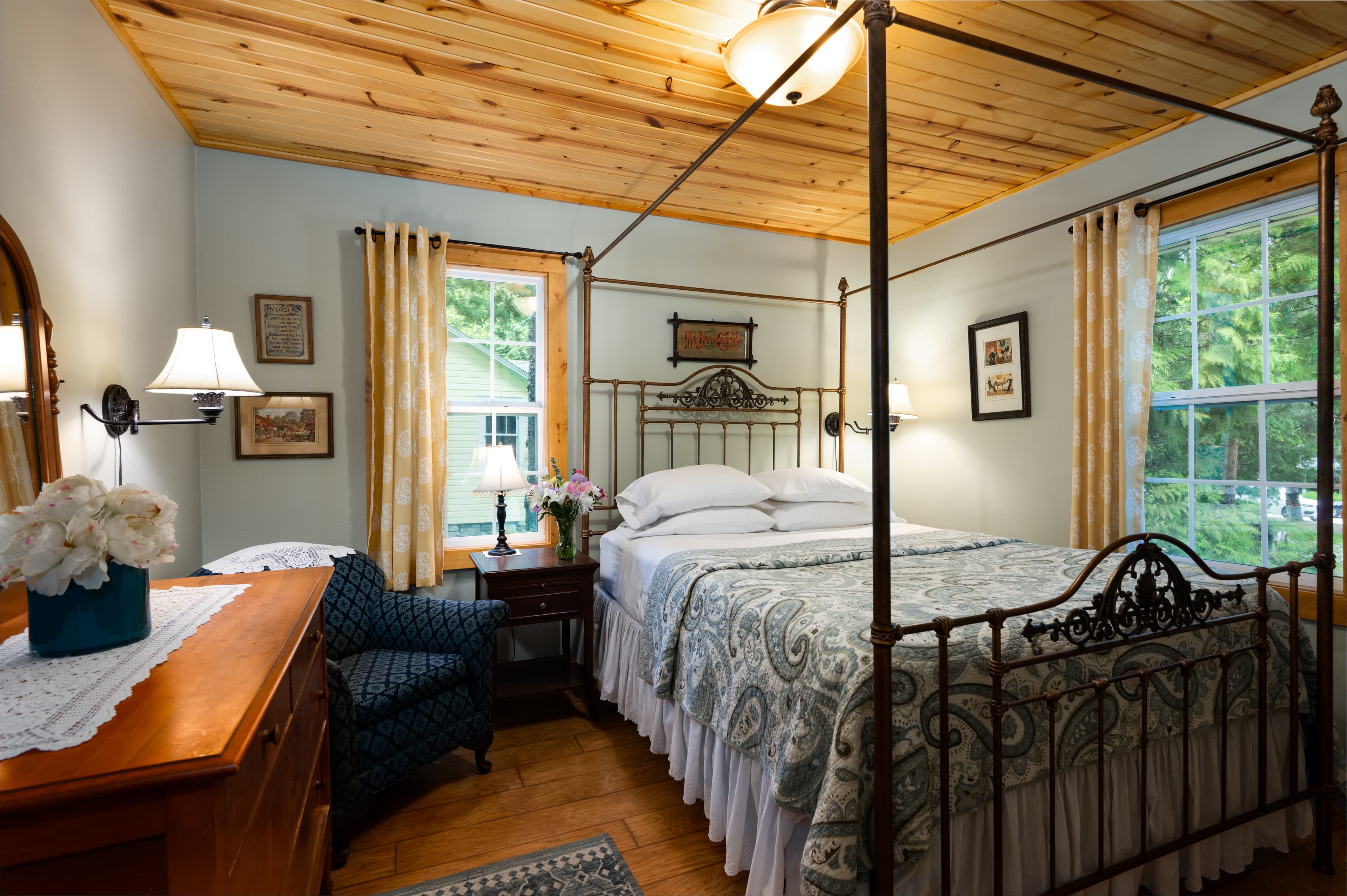 A warm and inviting bedroom with a wrought iron four-poster bed, light-colored walls, and a rustic wood-paneled ceiling.