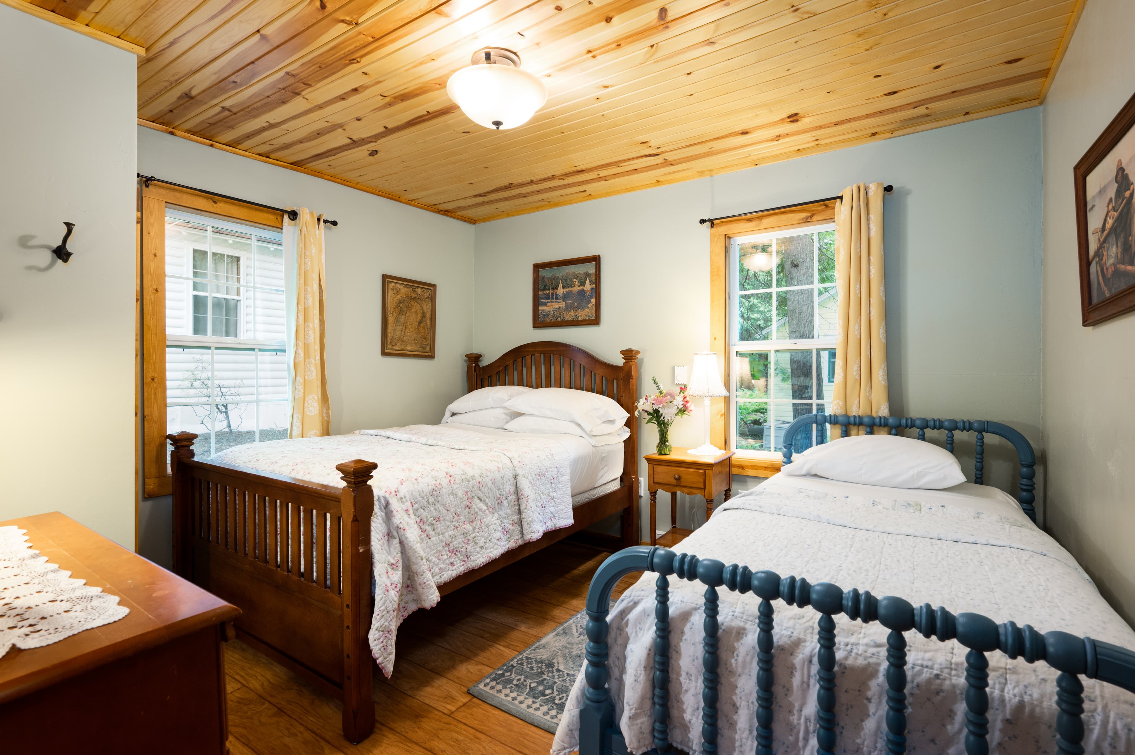 A quaint bedroom featuring two single beds, one with a wooden frame and the other with a blue metal frame, against light blue walls and a wood-paneled ceiling.