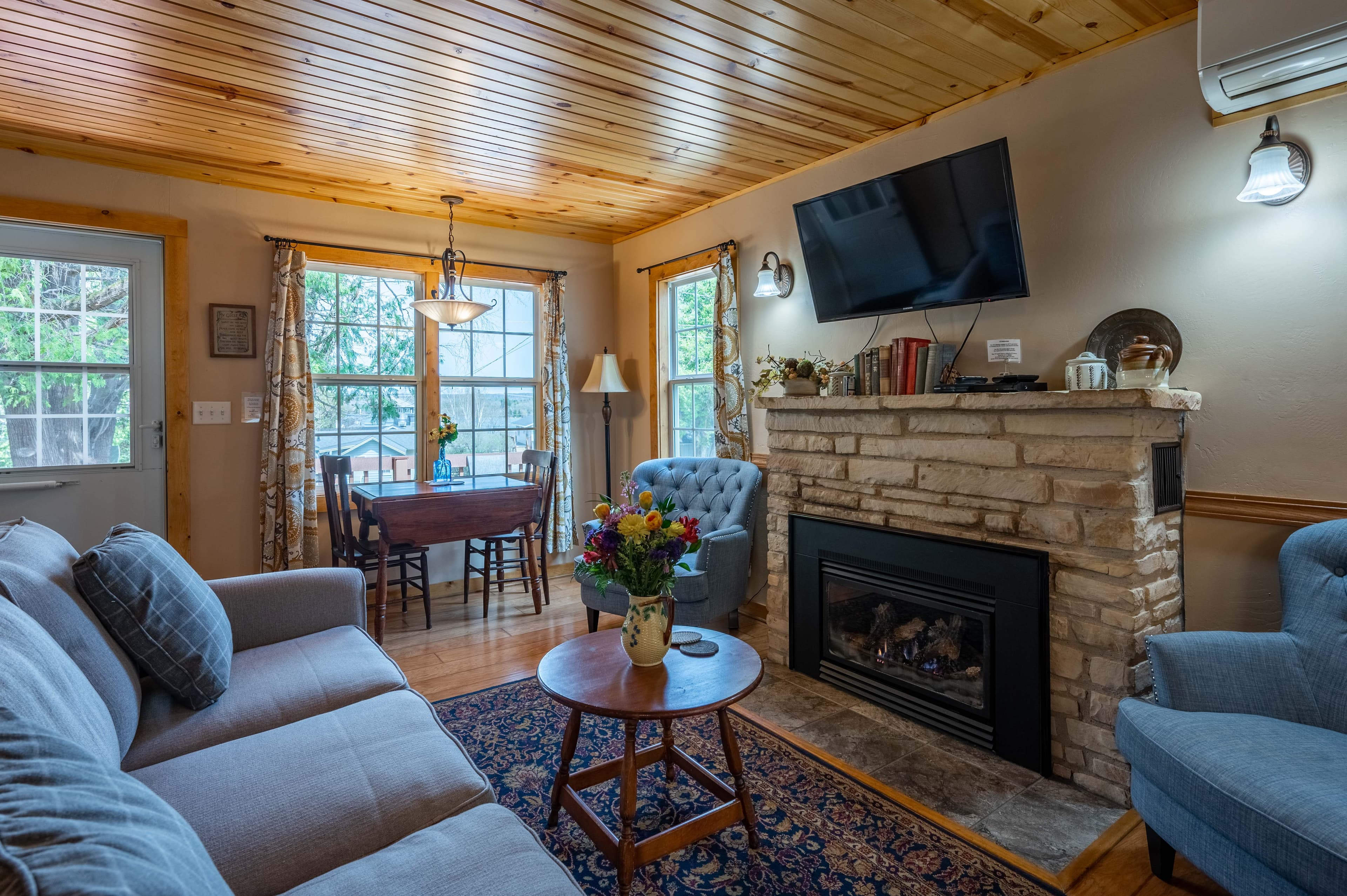 A welcoming living space with a stone fireplace, a wall-mounted TV, a comfy sofa, and a dining area, all under a warm wood-paneled ceiling.