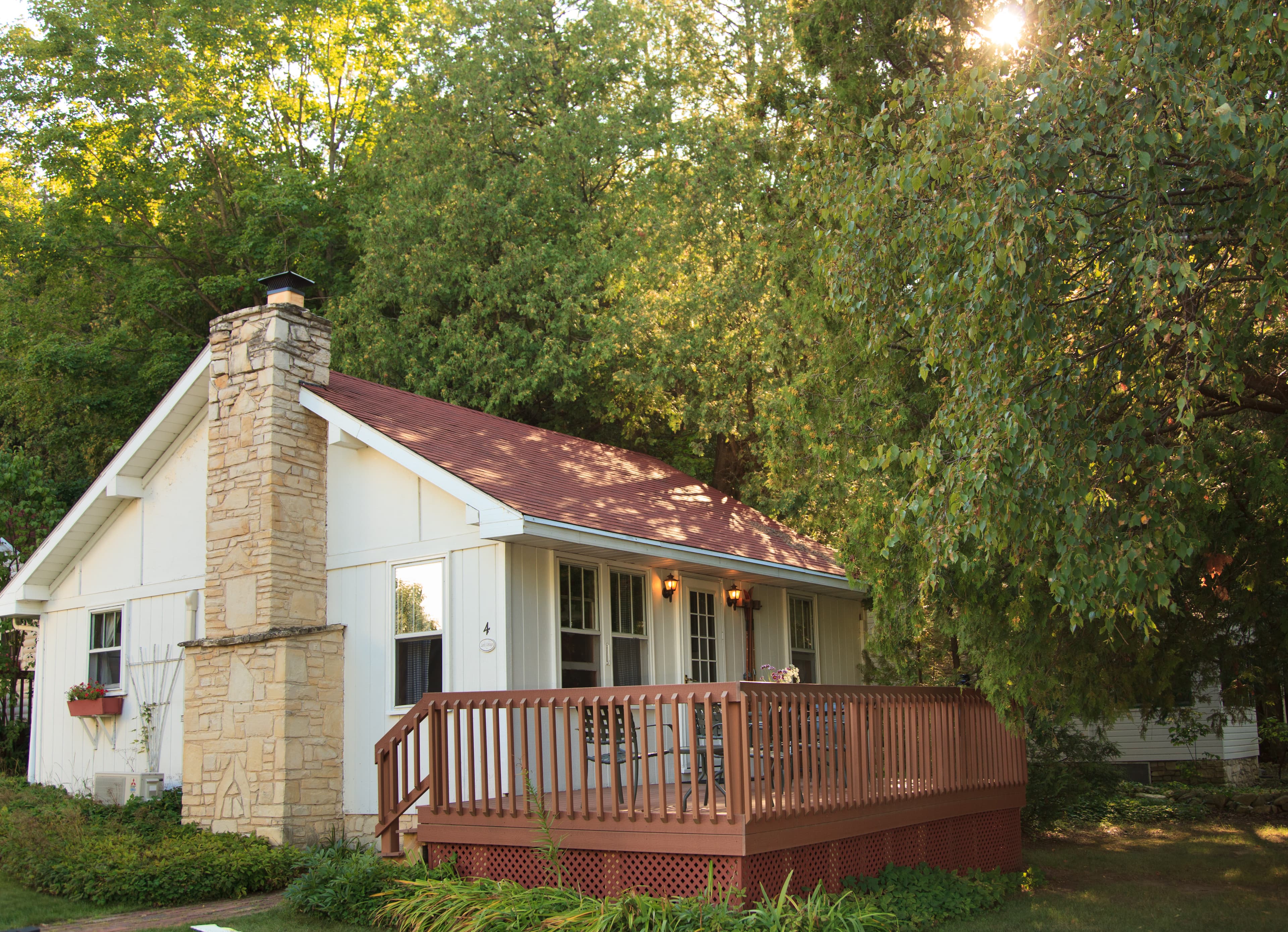 A quaint white cottage with a red roof, a prominent stone chimney, and a wooden deck, nestled among green trees with sunlight peeking through.