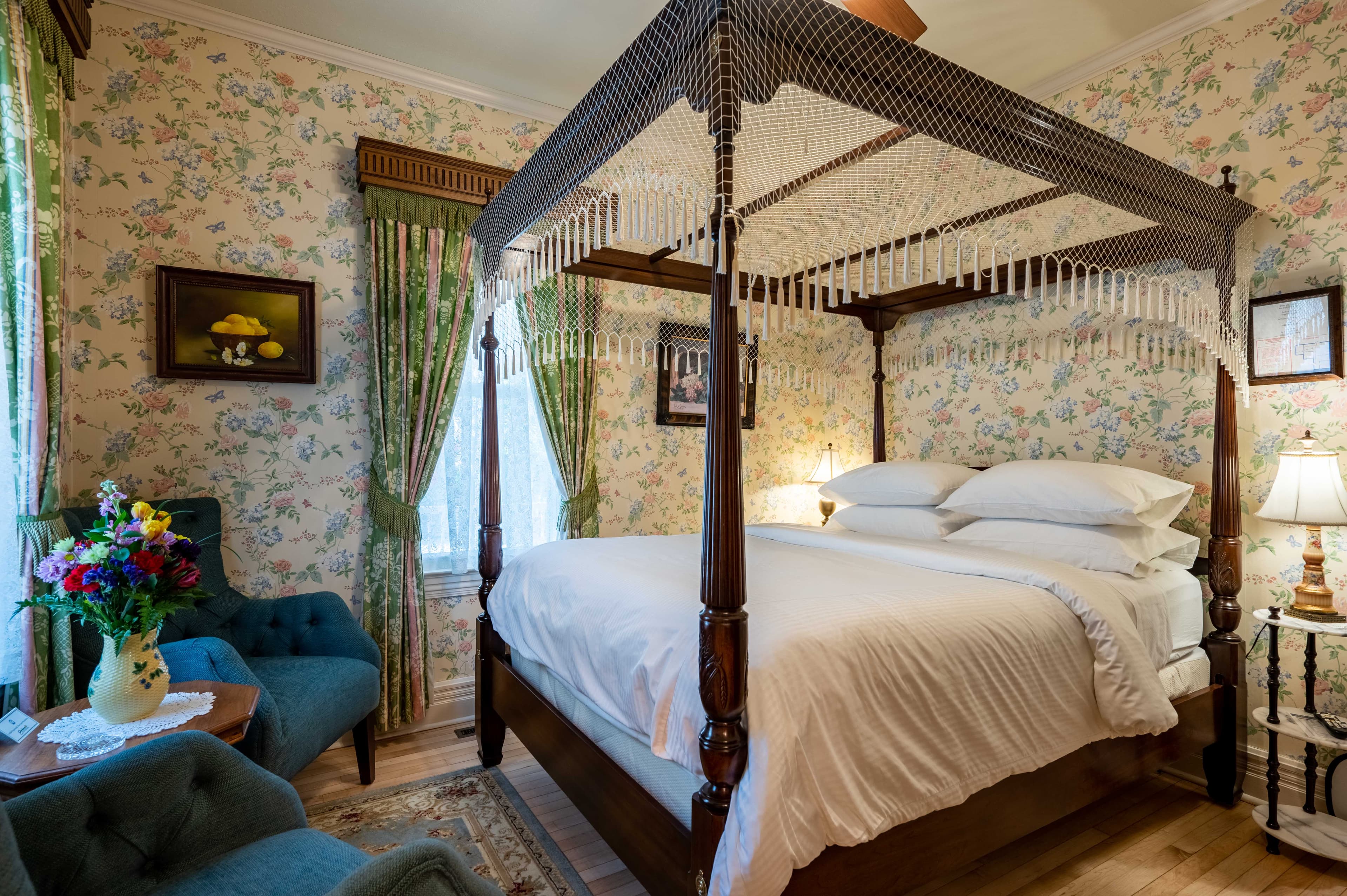 A classic bedroom featuring a dark wood four-poster bed, floral wallpaper, and two teal velvet chairs.