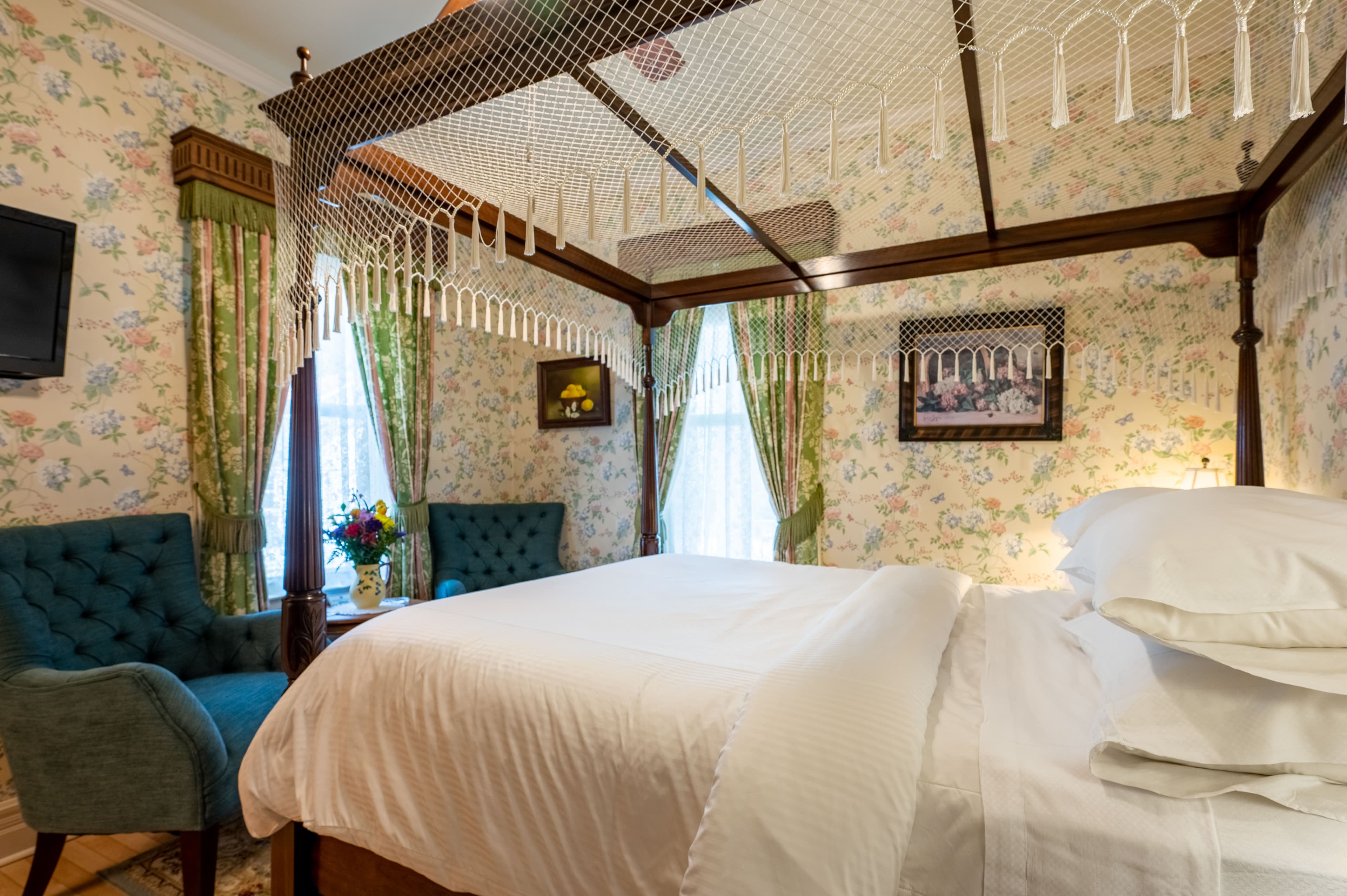A spacious bedroom featuring a dark wood four-poster bed with white bedding, floral wallpaper, and green patterned curtains.