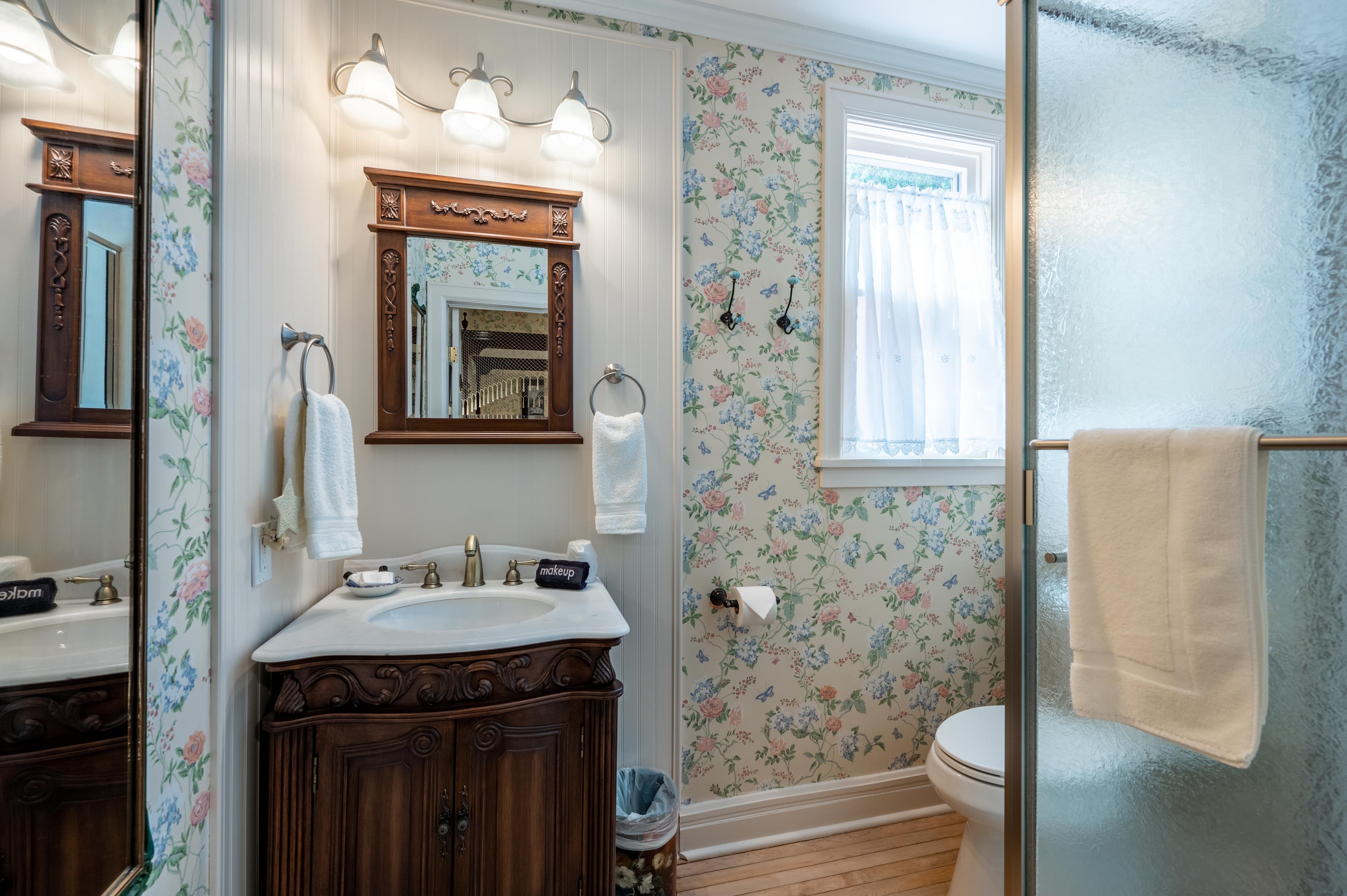 A well-lit bathroom with floral wallpaper, a wood vanity, a sink, and a glass shower enclosure.
