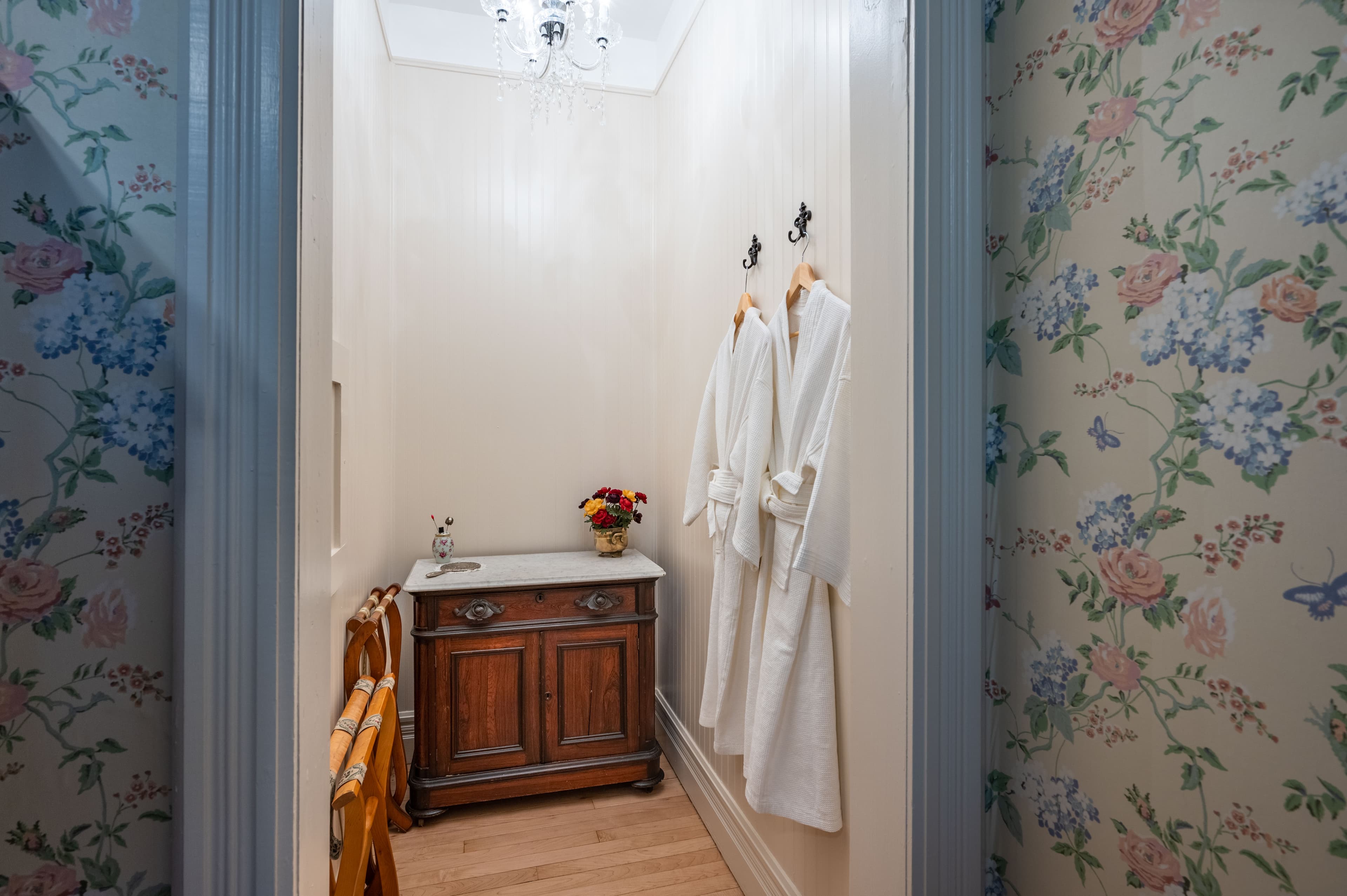 A hallway nook with floral wallpaper, a dark wood cabinet with a sink, and two white robes hanging on hooks.