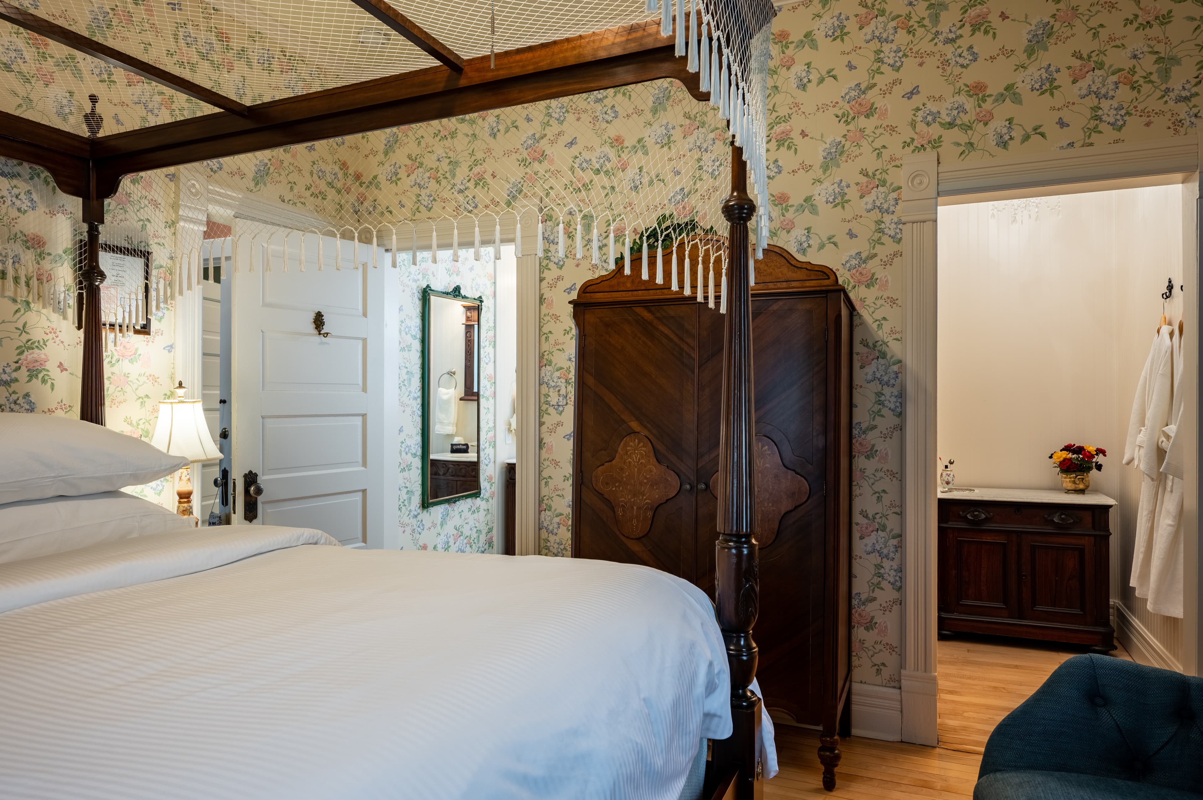 A charming bedroom with floral wallpaper, a dark wood four-poster queen bed, and a wardrobe, with a partial view of an adjoining nook.