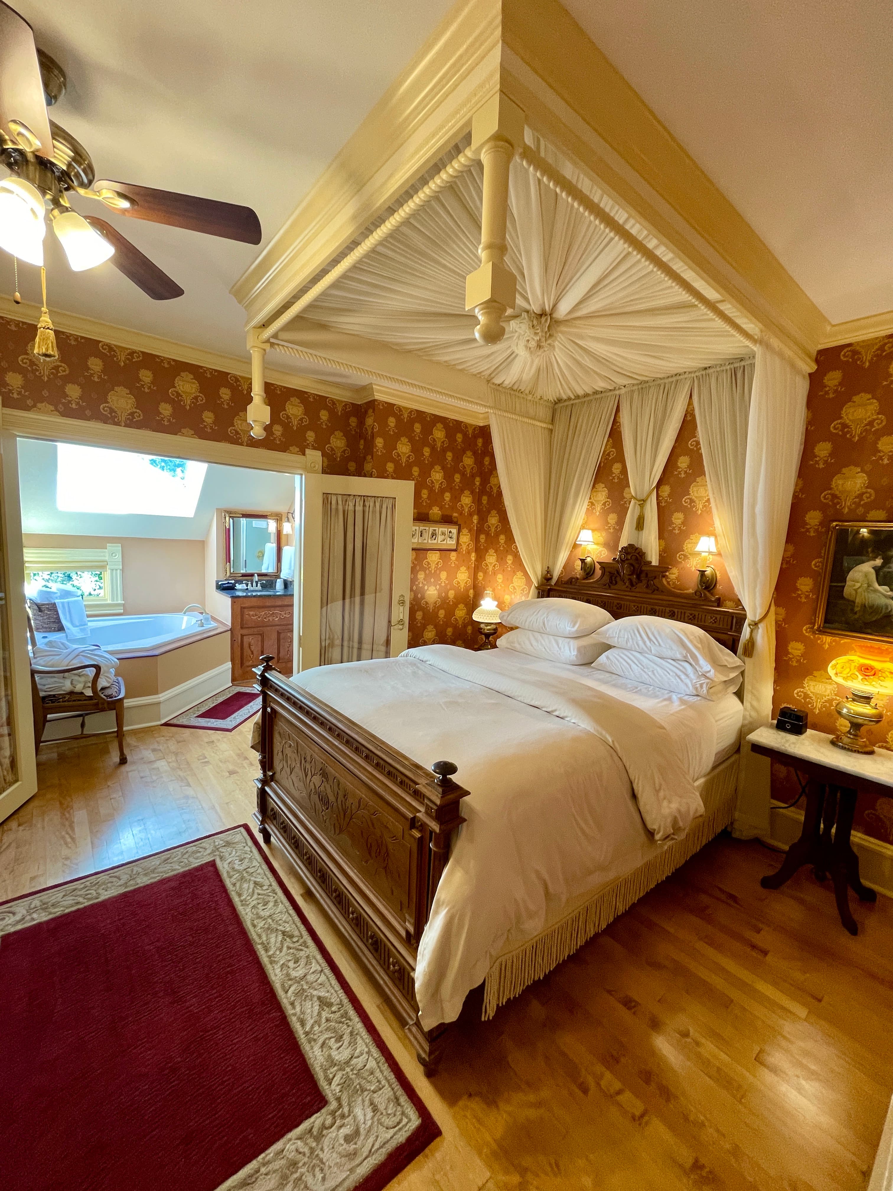 A luxurious bedroom featuring a four-poster bed with canopy, gold and red wallpaper, and an adjoining bathroom with a spa tub.