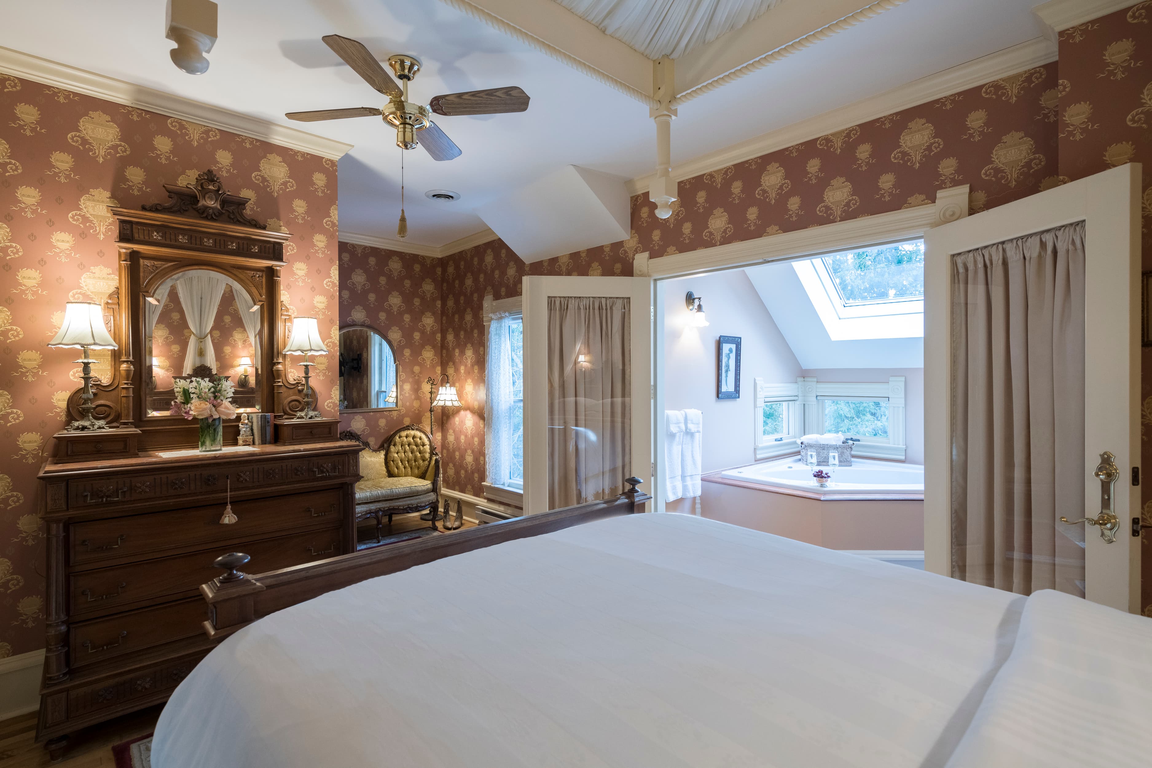 A bedroom with ornate dark wood furniture, gold and red wallpaper, a ceiling fan, and an open doorway to a bathroom with a tub.