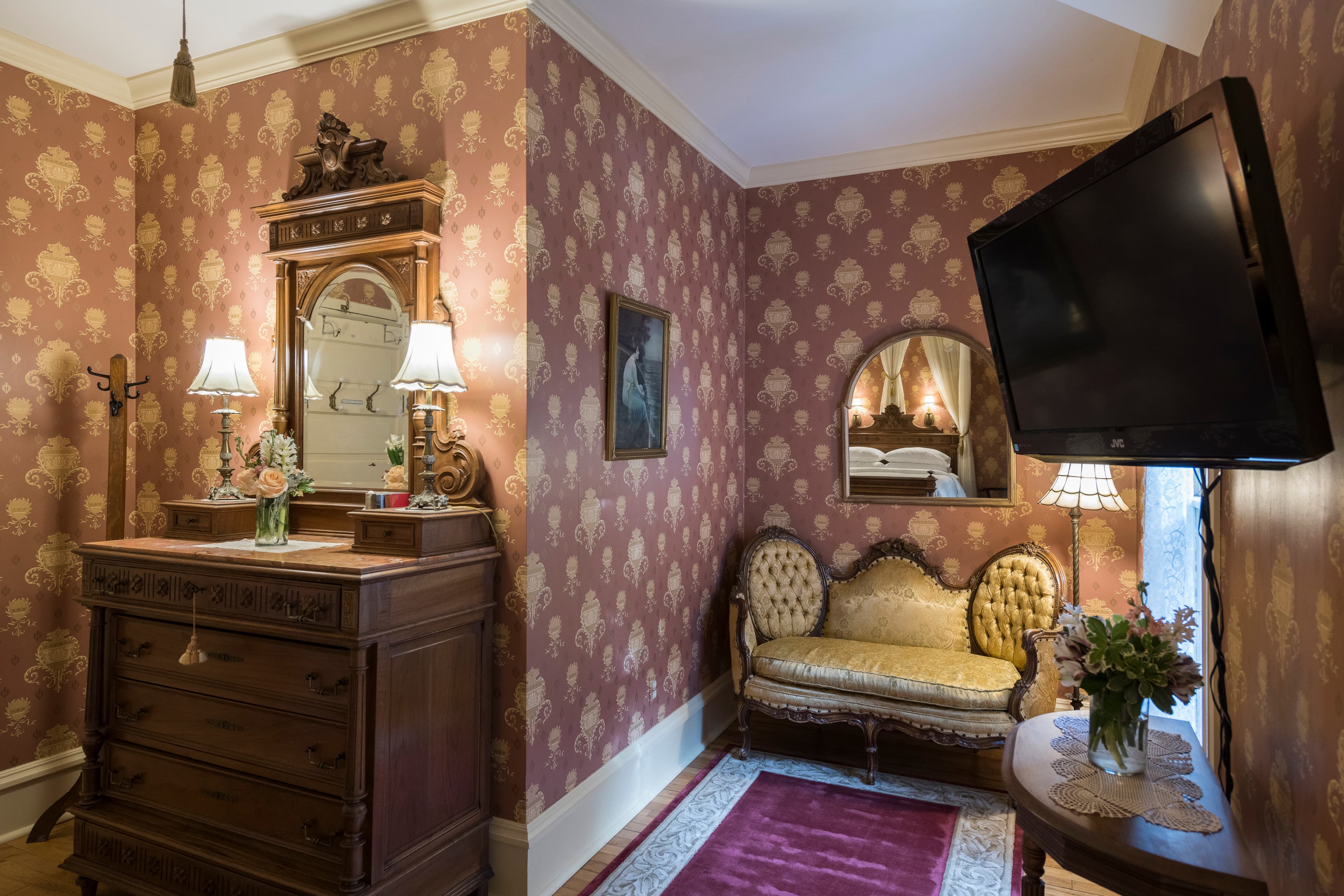 A richly decorated sitting area with red and gold patterned wallpaper, a wooden dresser with a mirror, and a small chaise lounge.