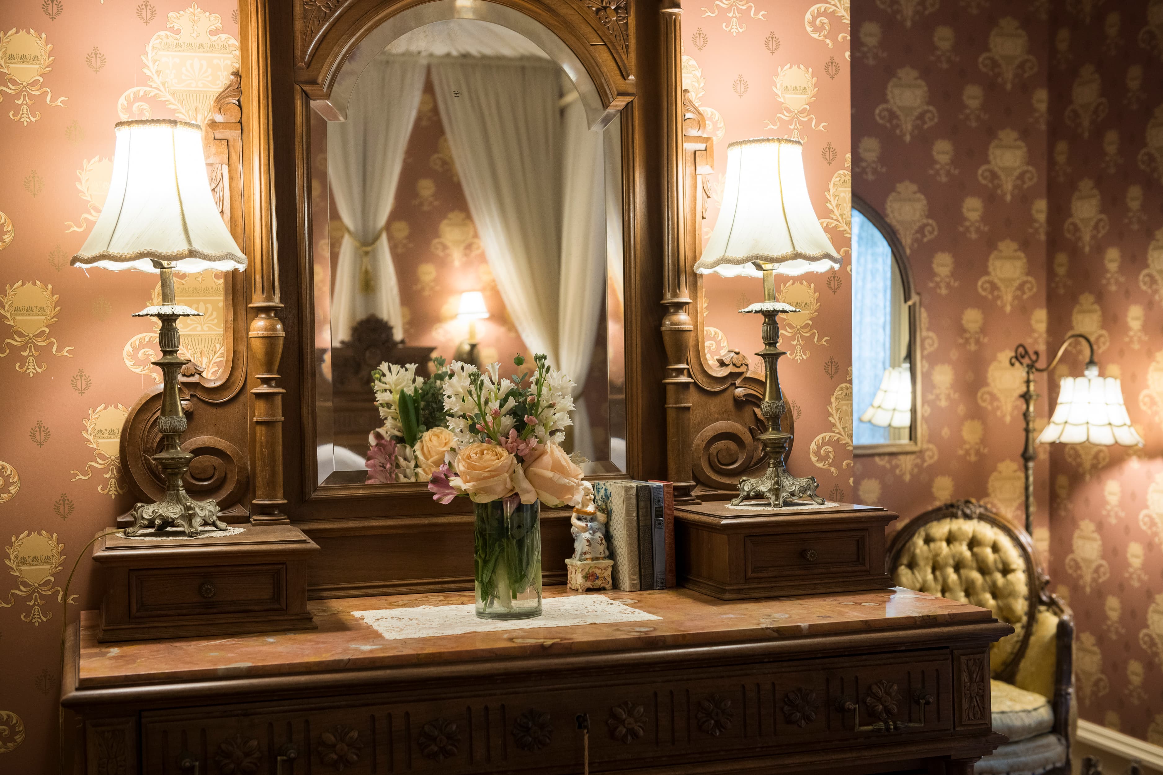An antique wooden vanity with an ornate mirror, two lamps, and a vase of flowers, set against a red and gold patterned wallpaper.