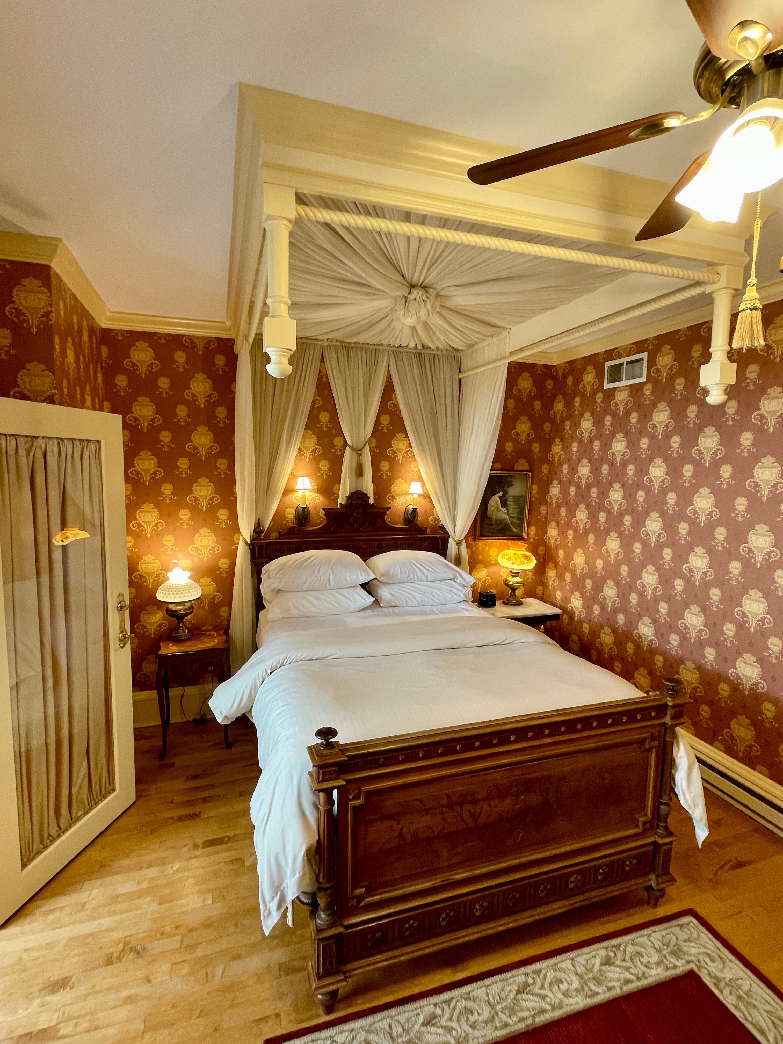 A bedroom with a richly carved wooden four-poster bed with white canopy, gold and red patterned wallpaper, and a ceiling fan.