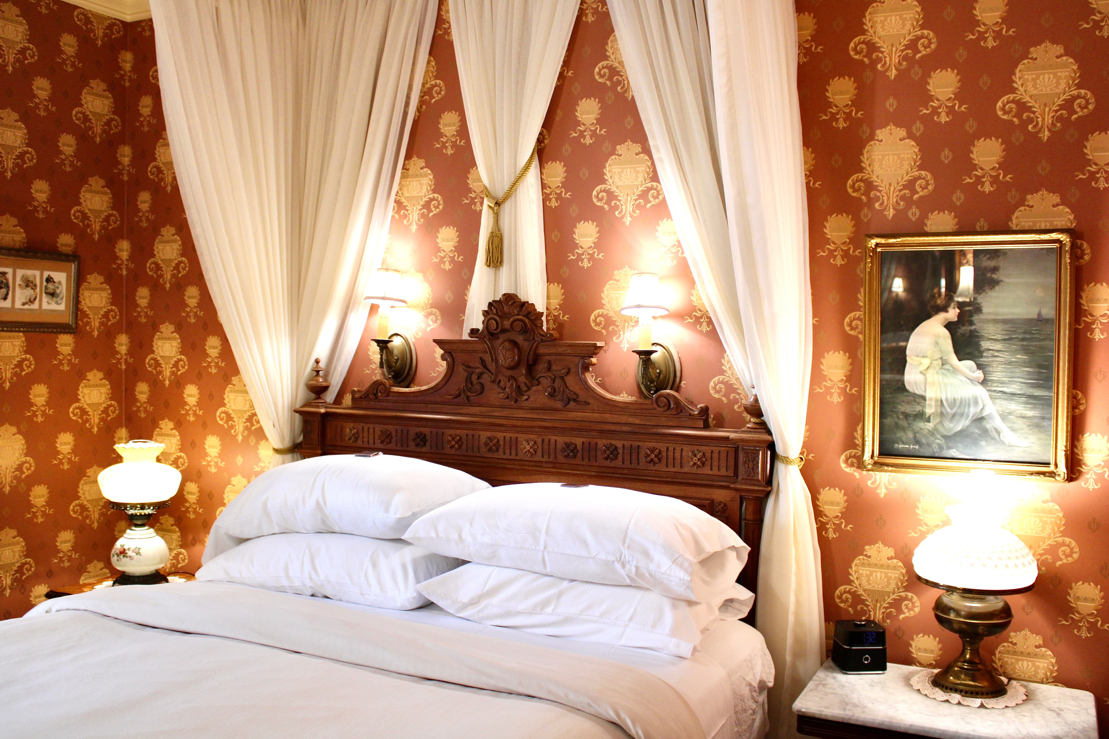 A bedroom with a richly carved wooden four-poster bed with white canopy, gold and red patterned wallpaper, and a ceiling fan.