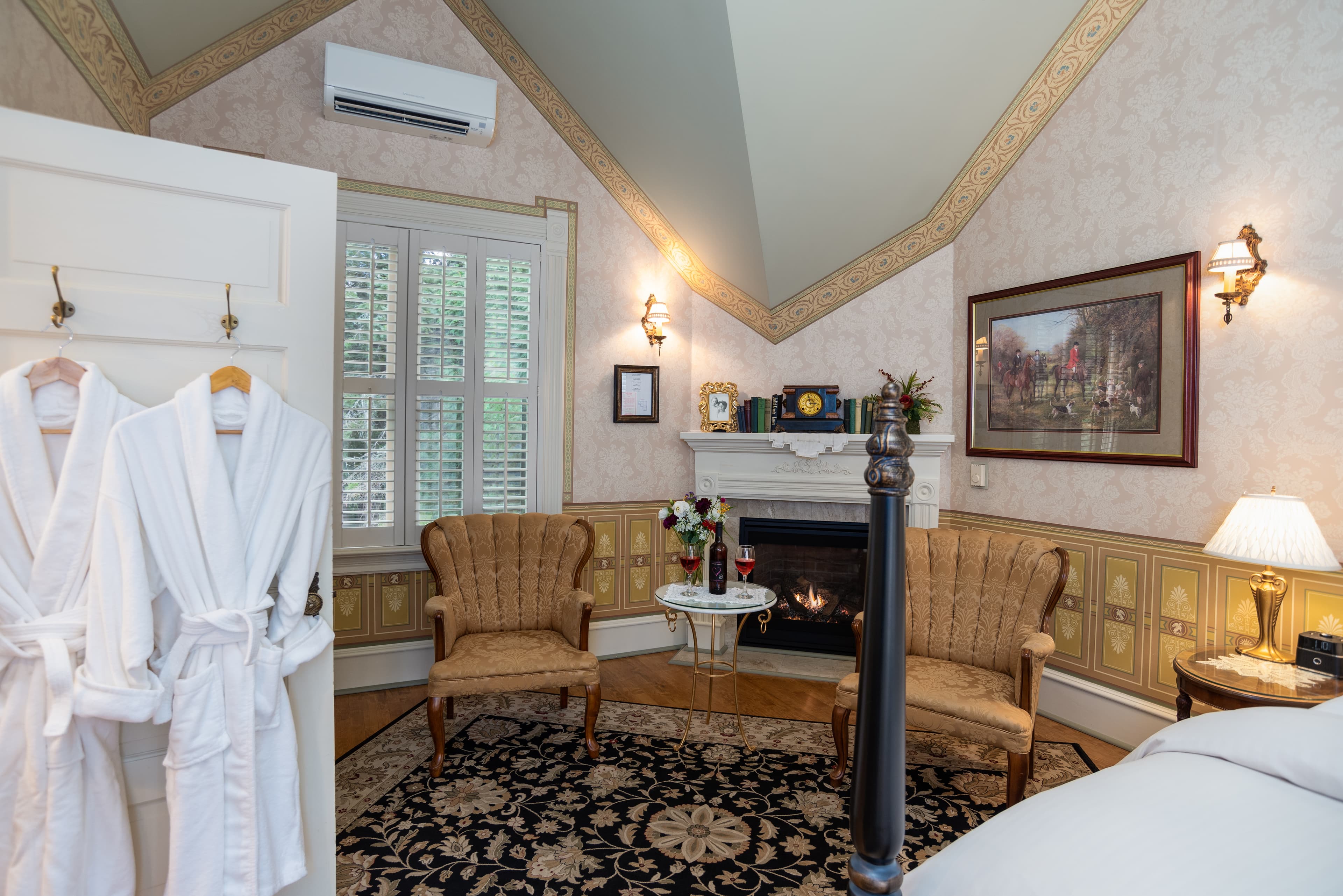 A charming bedroom featuring a fireplace, two armchairs, a large window, and two white robes hanging on a door, with an elegant ambiance.