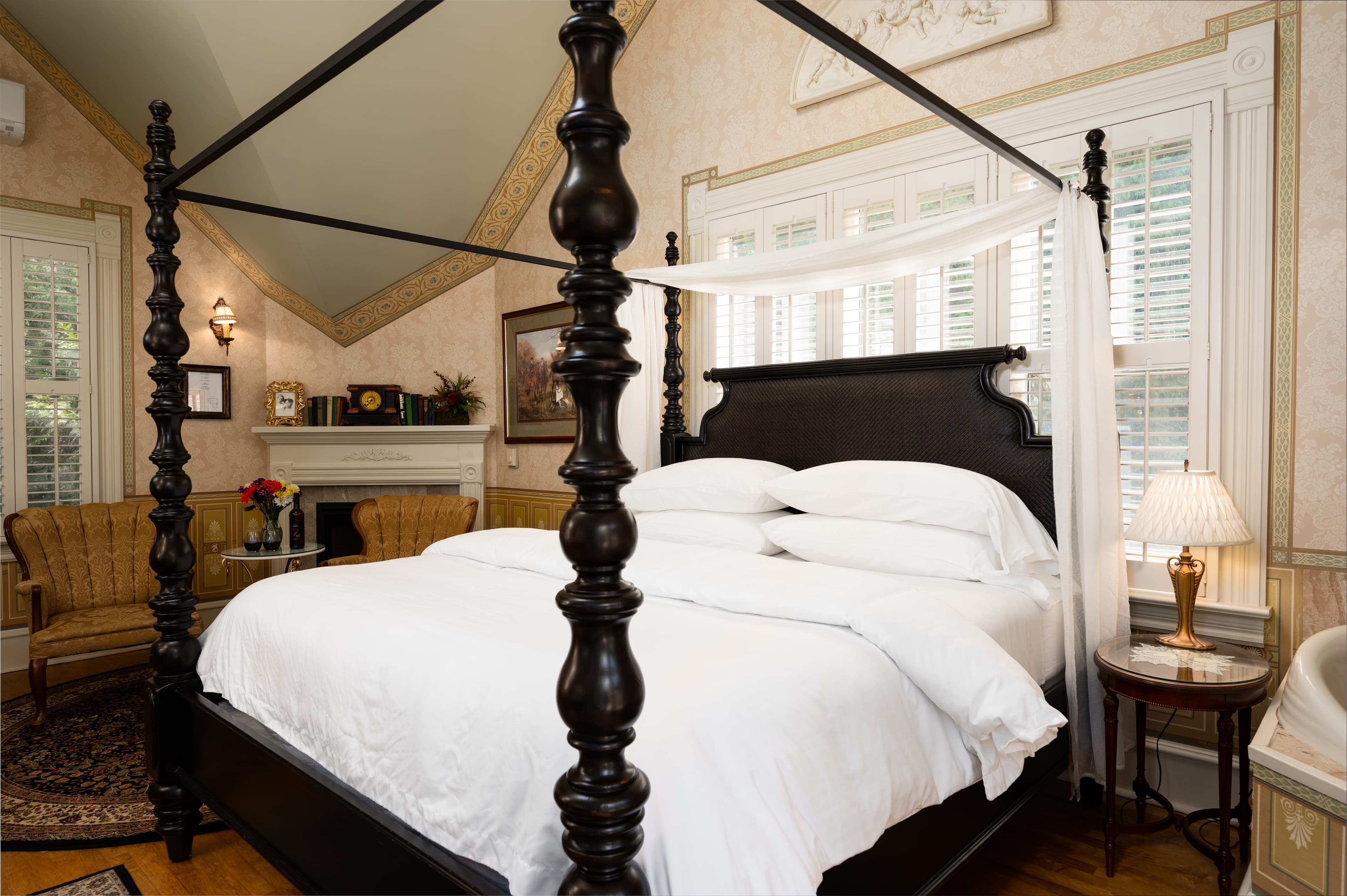 A classic bedroom featuring a dark wood four-poster bed with white bedding, a fireplace, and two armchairs.