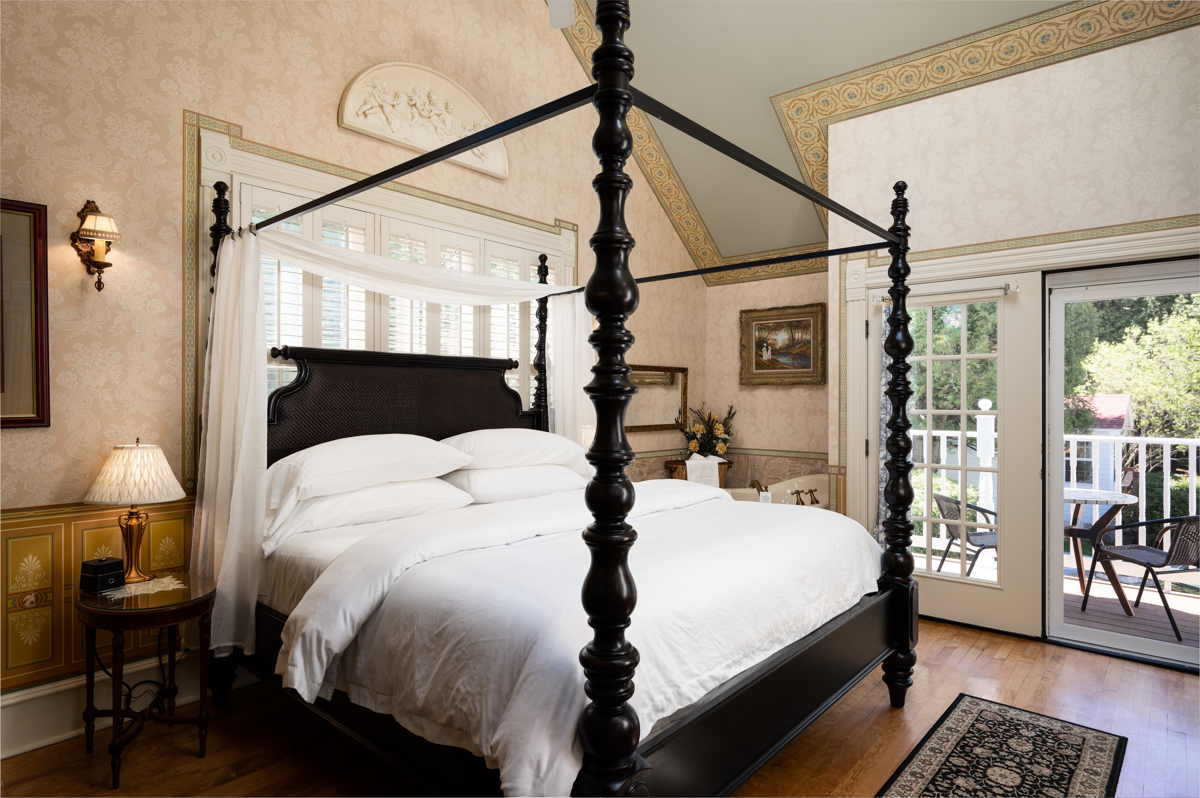 A stately bedroom featuring a dark wood four-poster bed with white bedding, ornate wallpaper, and French doors leading to a bright balcony.