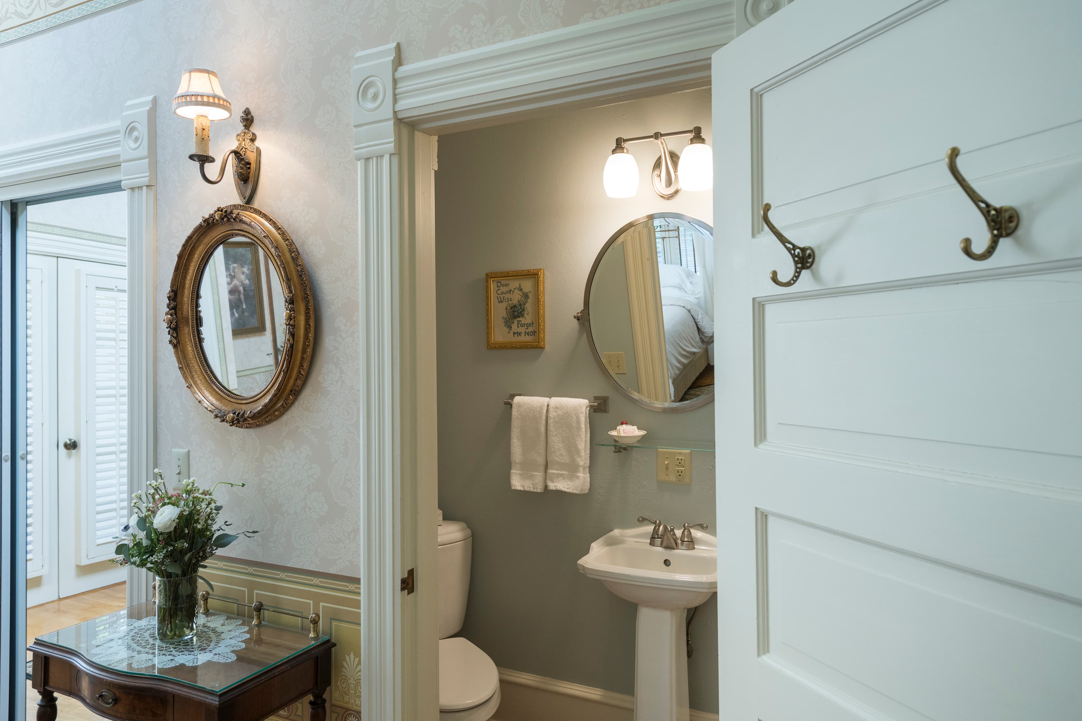 A bright entryway leading to a bathroom with a pedestal sink, toilet, and round mirror, featuring elegant decor and a classic ambiance.