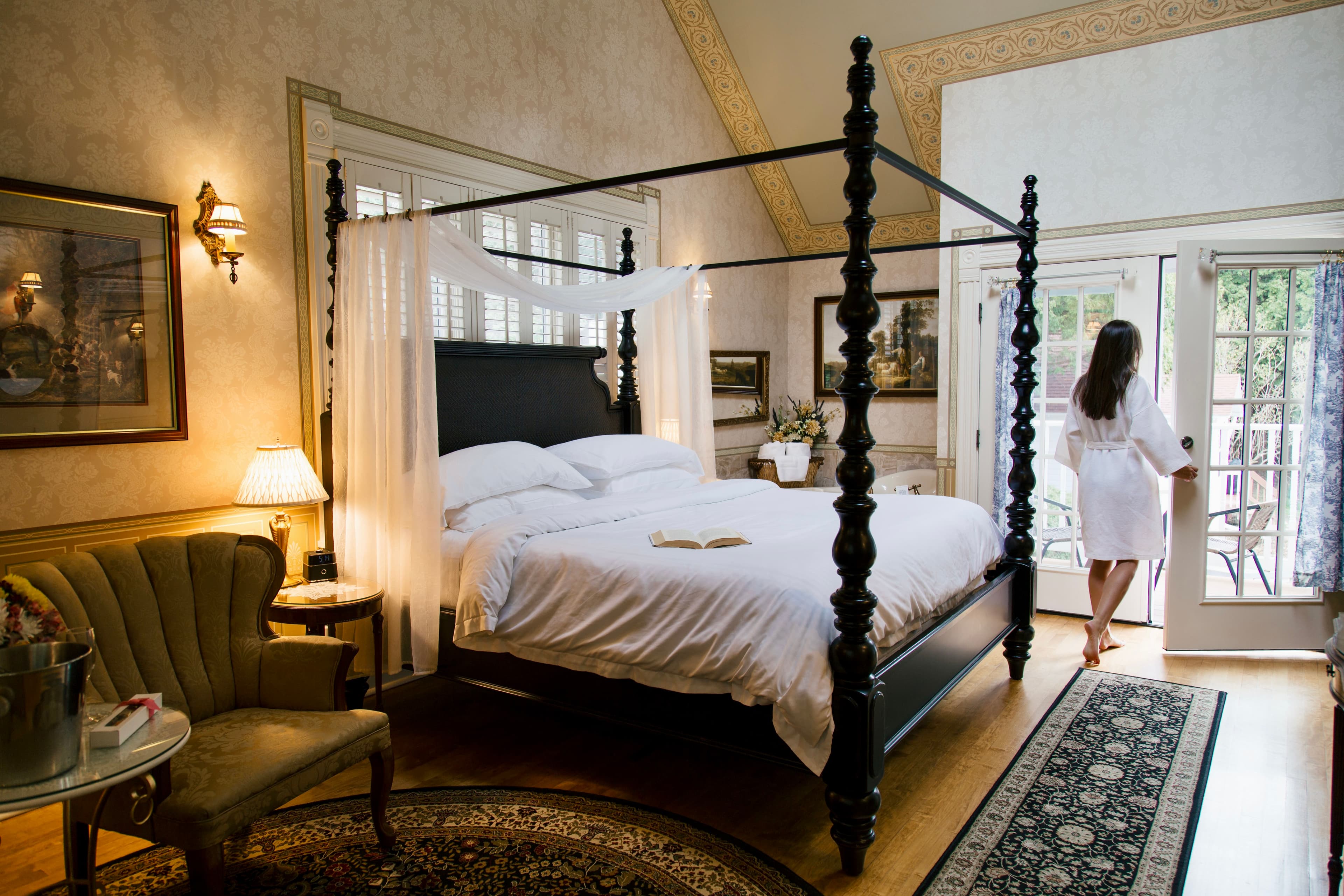A serene bedroom with a dark wood four-poster bed, a sitting area, and a person in a white robe opening French doors to the outside.
