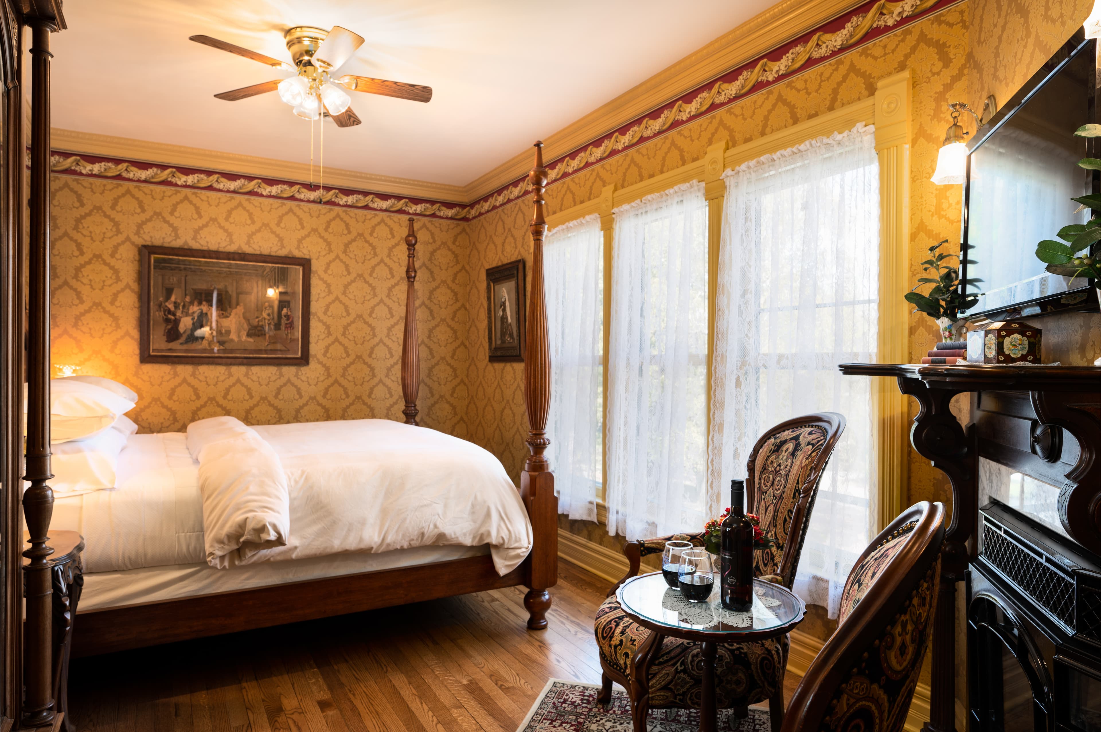 A grand bedroom featuring a wooden four-poster bed, ornate gold and red wallpaper, a sitting area, and a large window.