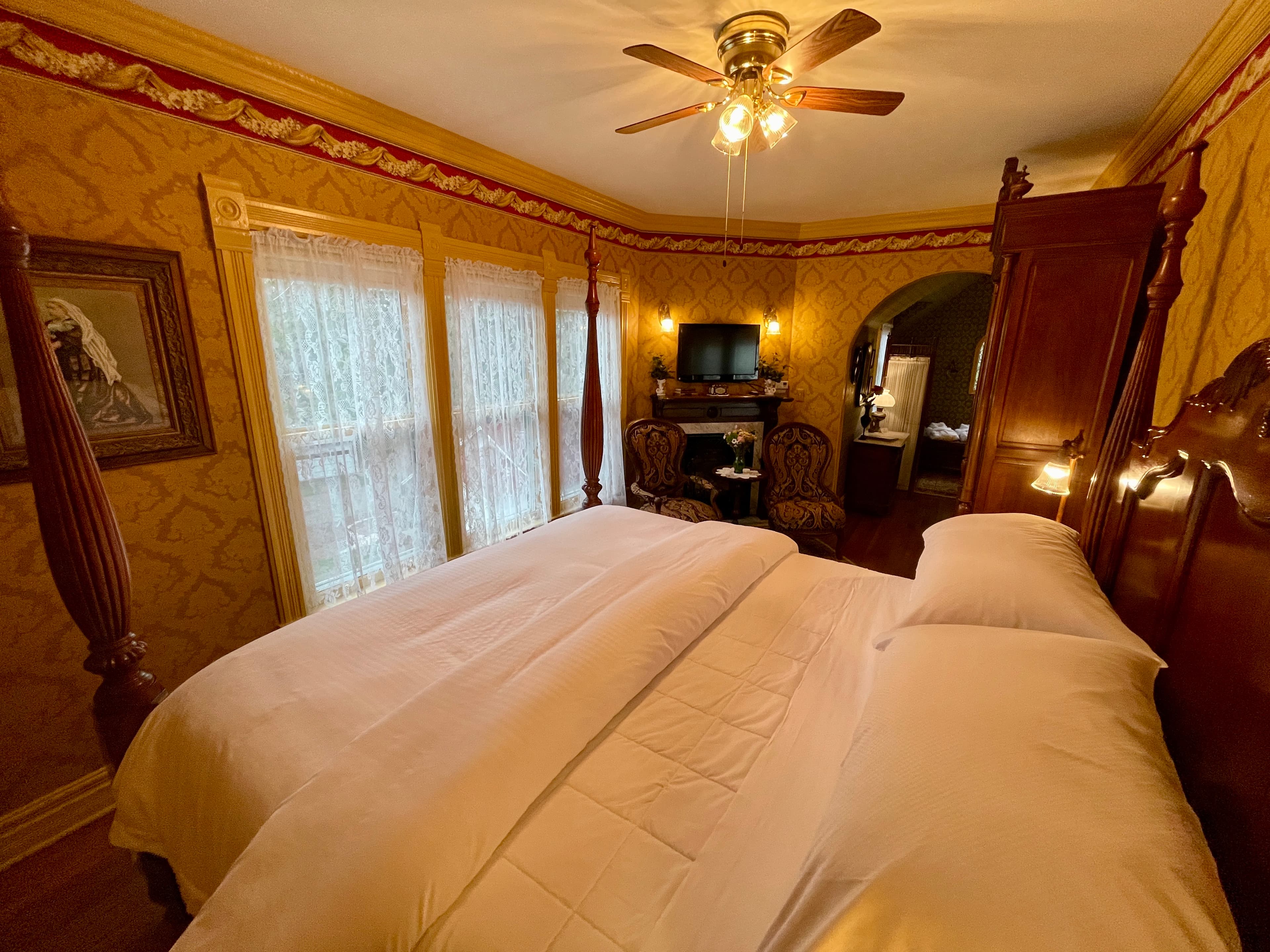 A lavish bedroom featuring a large four-poster bed with white bedding, ornate gold and red wallpaper, and a ceiling fan.