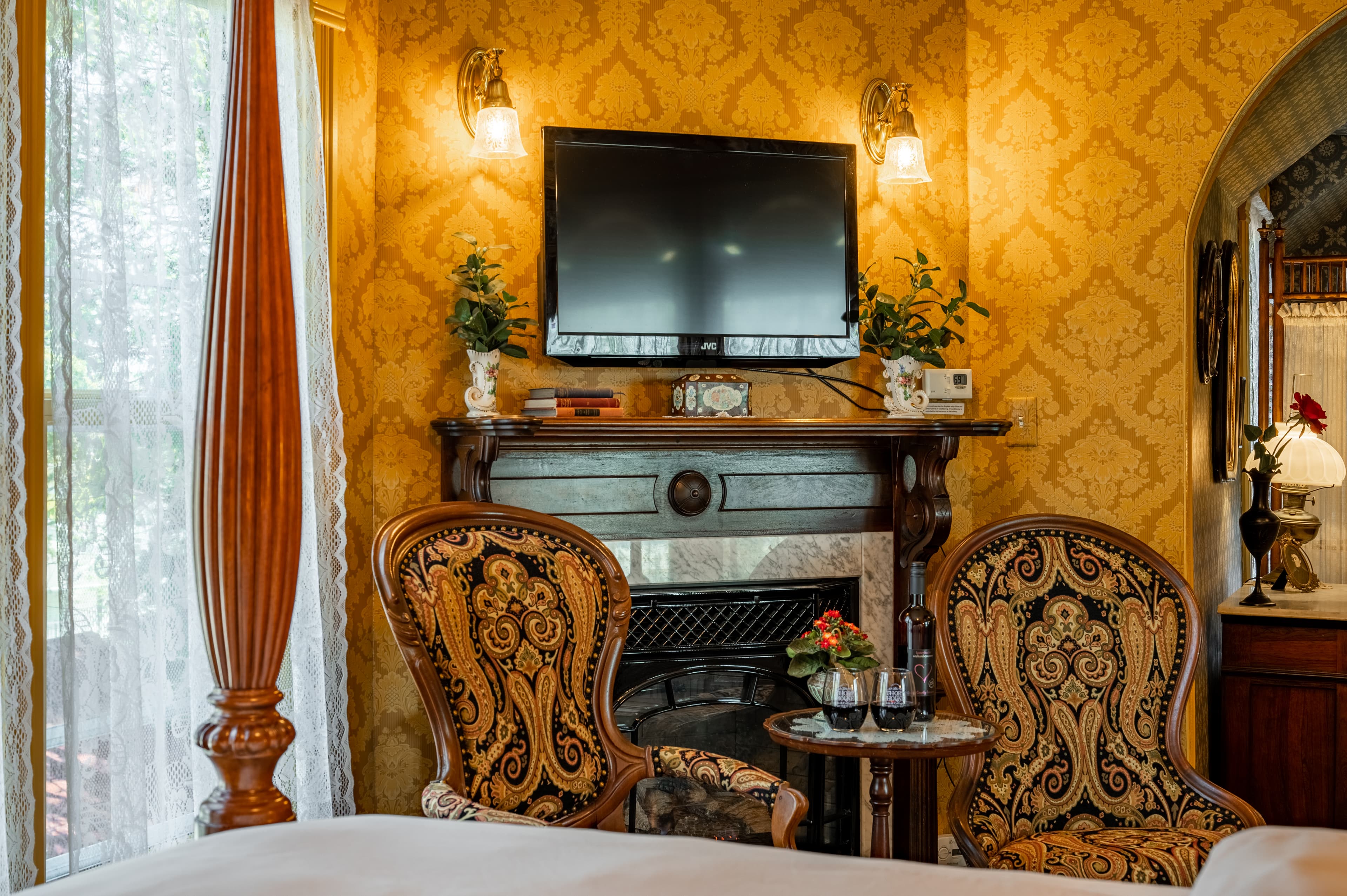 A richly decorated bedroom with a fireplace, two ornate chairs, a mounted television, and gold patterned wallpaper.
