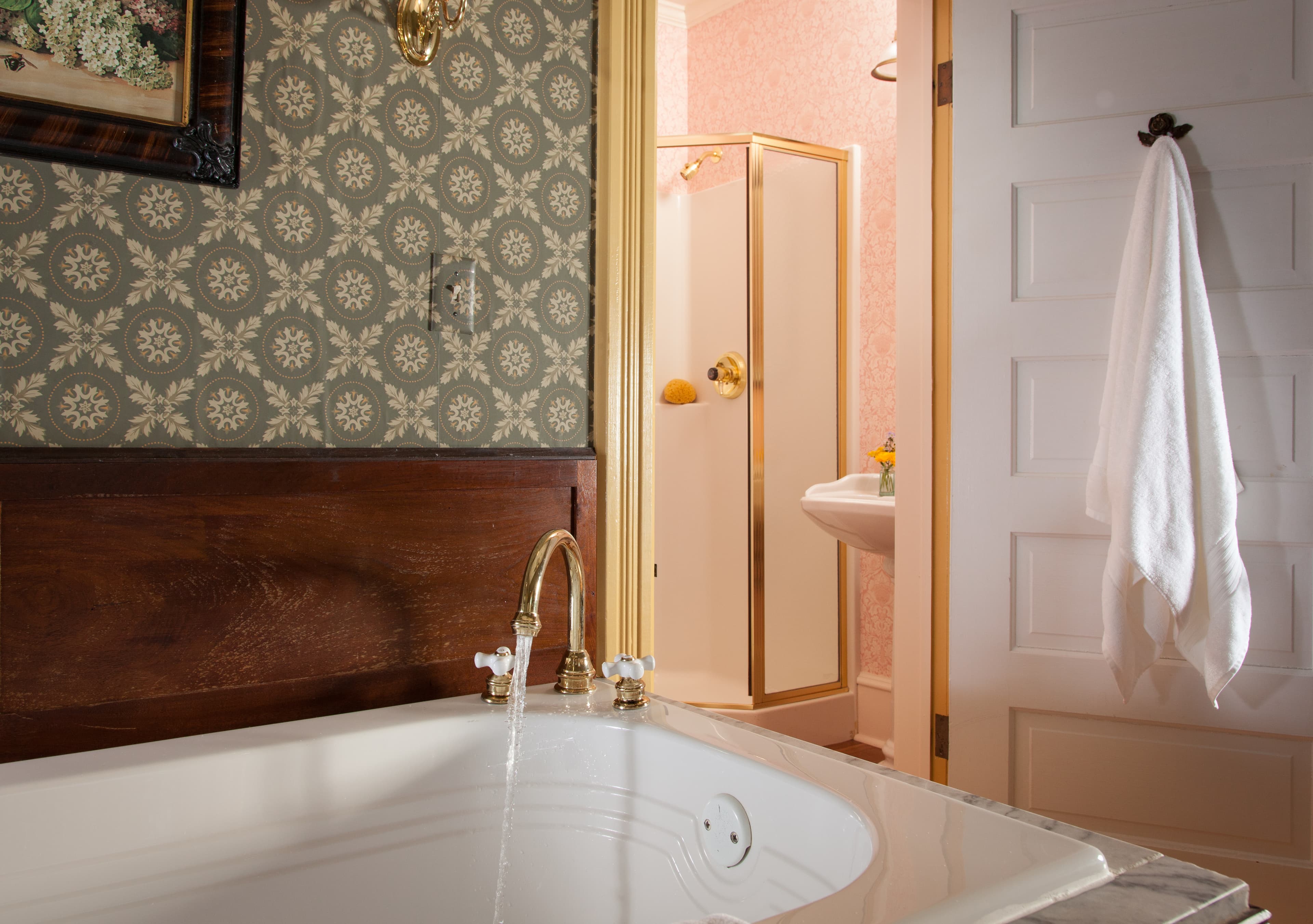 A luxurious bathroom featuring a large soaking tub with brass fixtures, patterned wallpaper, and a peek into an adjoining shower and sink area.