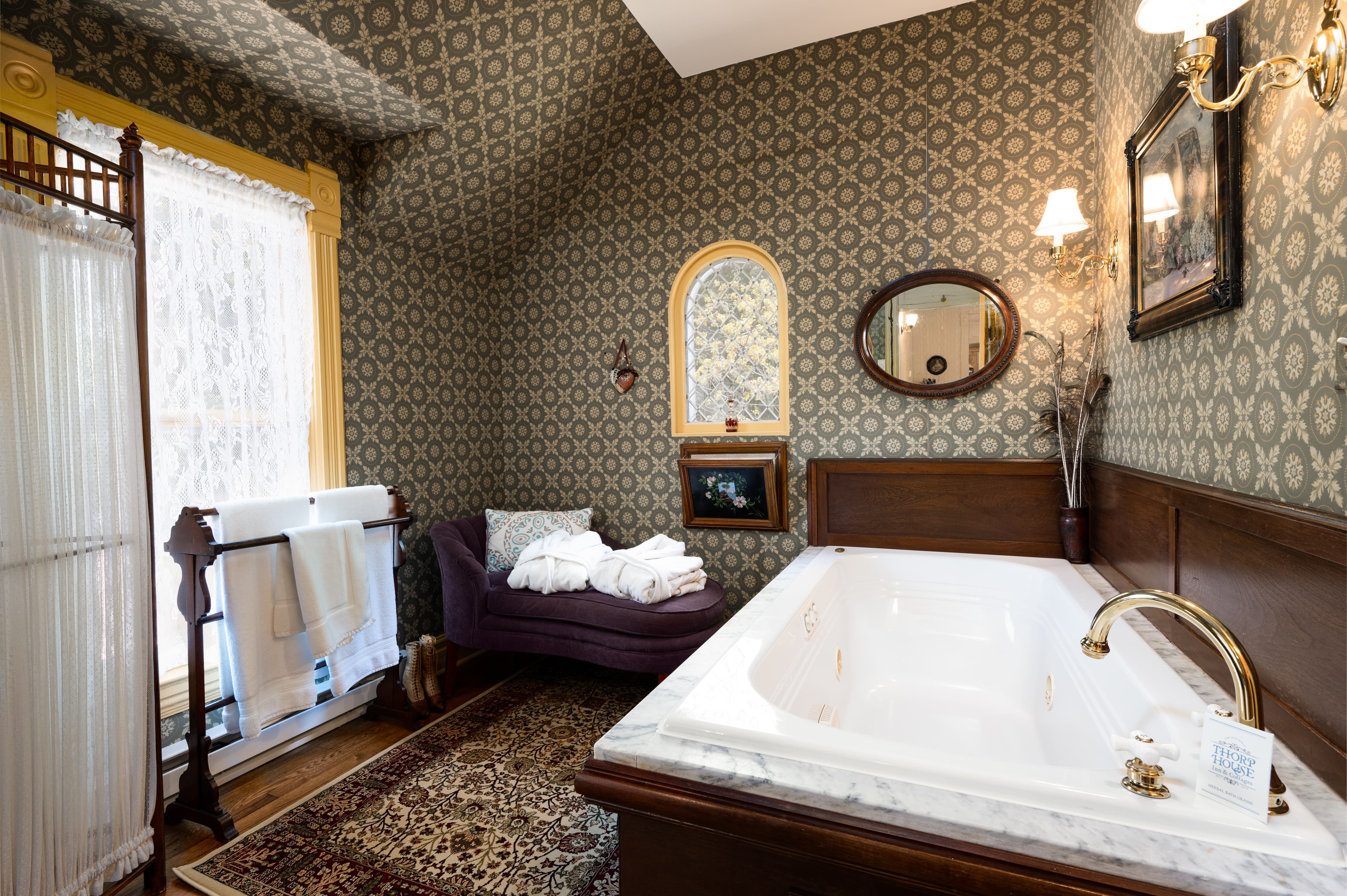 A grand bathroom featuring a large soaking tub, patterned wallpaper, a chaise lounge, and elegant decor, creating a luxurious retreat.