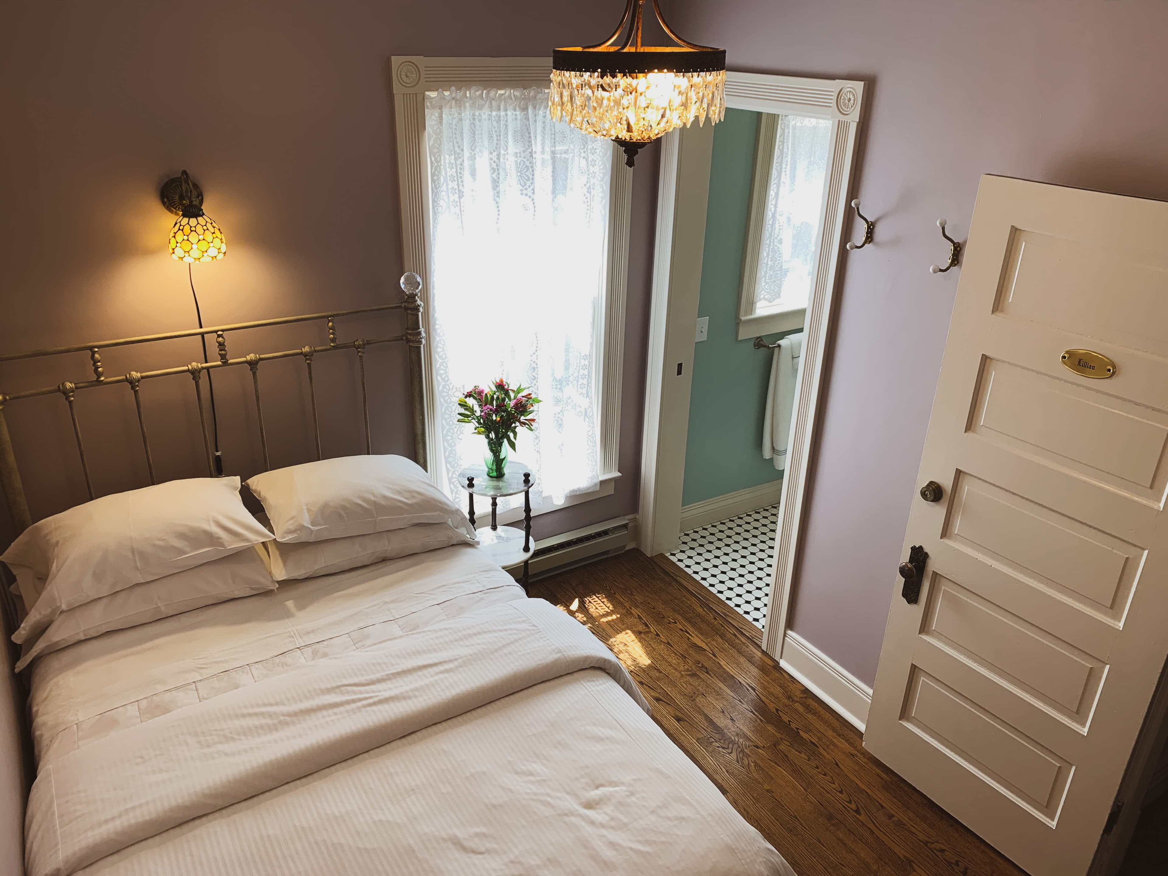 A charming bedroom with lavender walls, a bed with white bedding, a bright doorway leading to a bathroom, and hardwood floors.