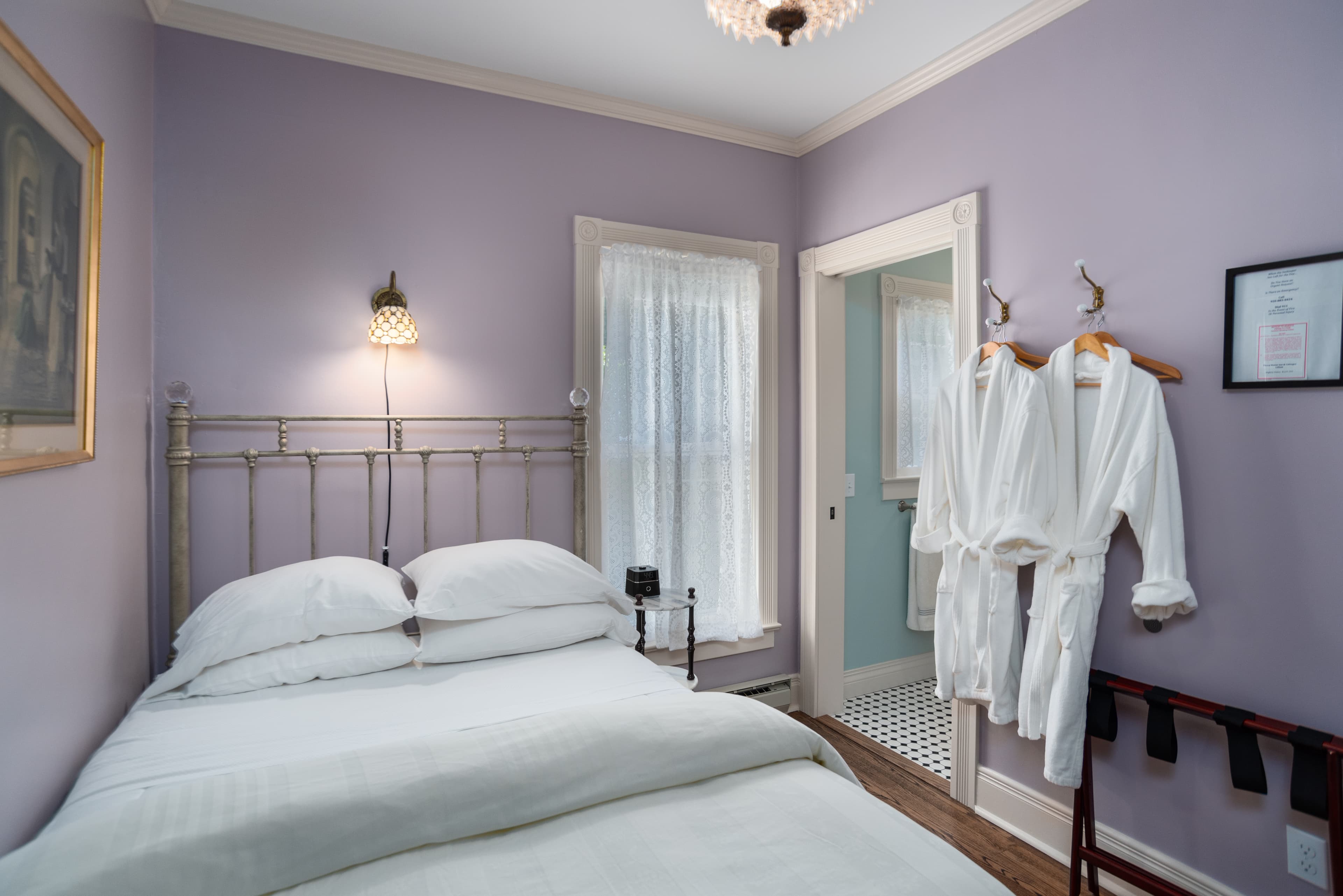 A cozy bedroom with lavender walls, a white bed, and two white robes hanging near the doorway to a bathroom.