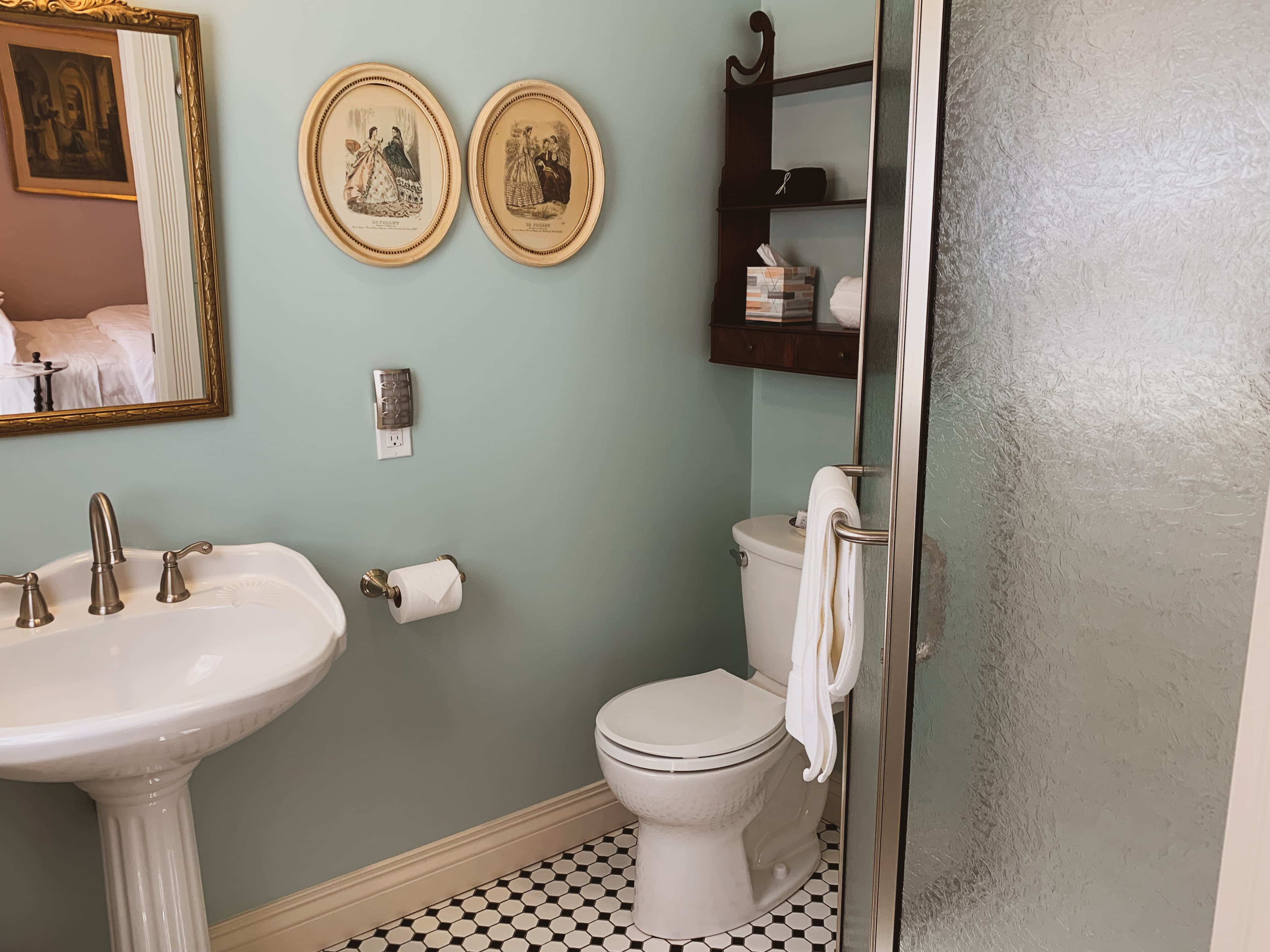 A bright bathroom with light blue walls, a pedestal sink, a toilet, a shower, and black and white checkered floor tiles.