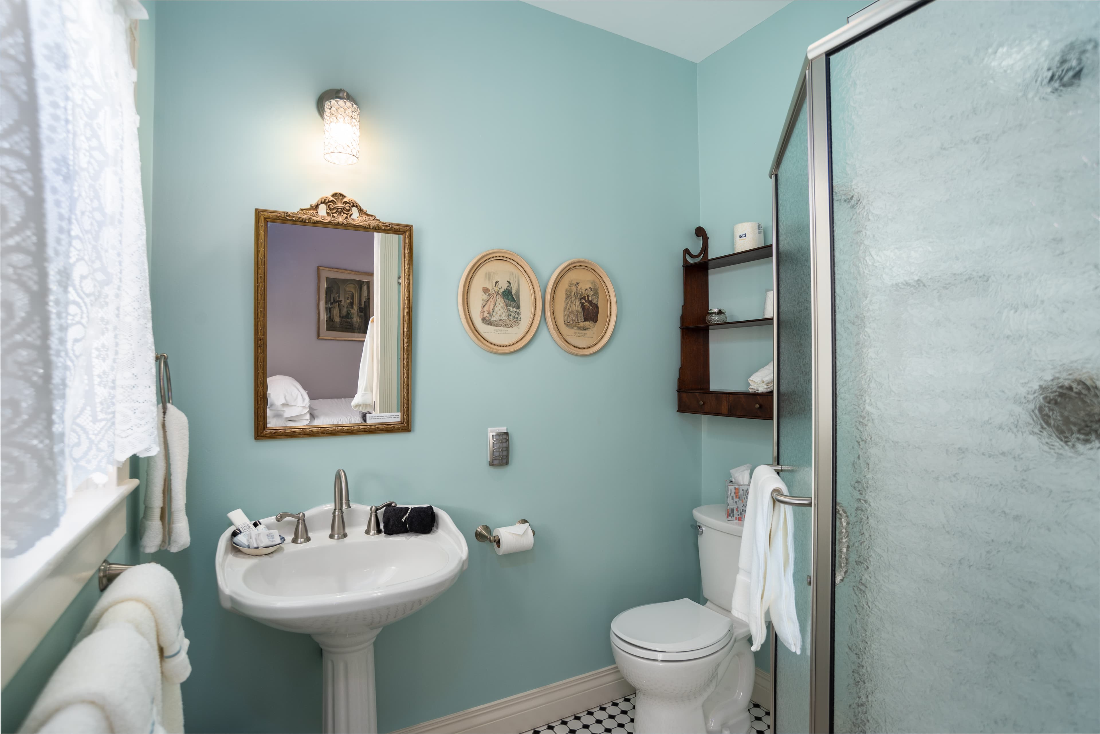 A bright bathroom with light blue walls, a pedestal sink, a toilet, a shower, and black and white checkered floor tiles.