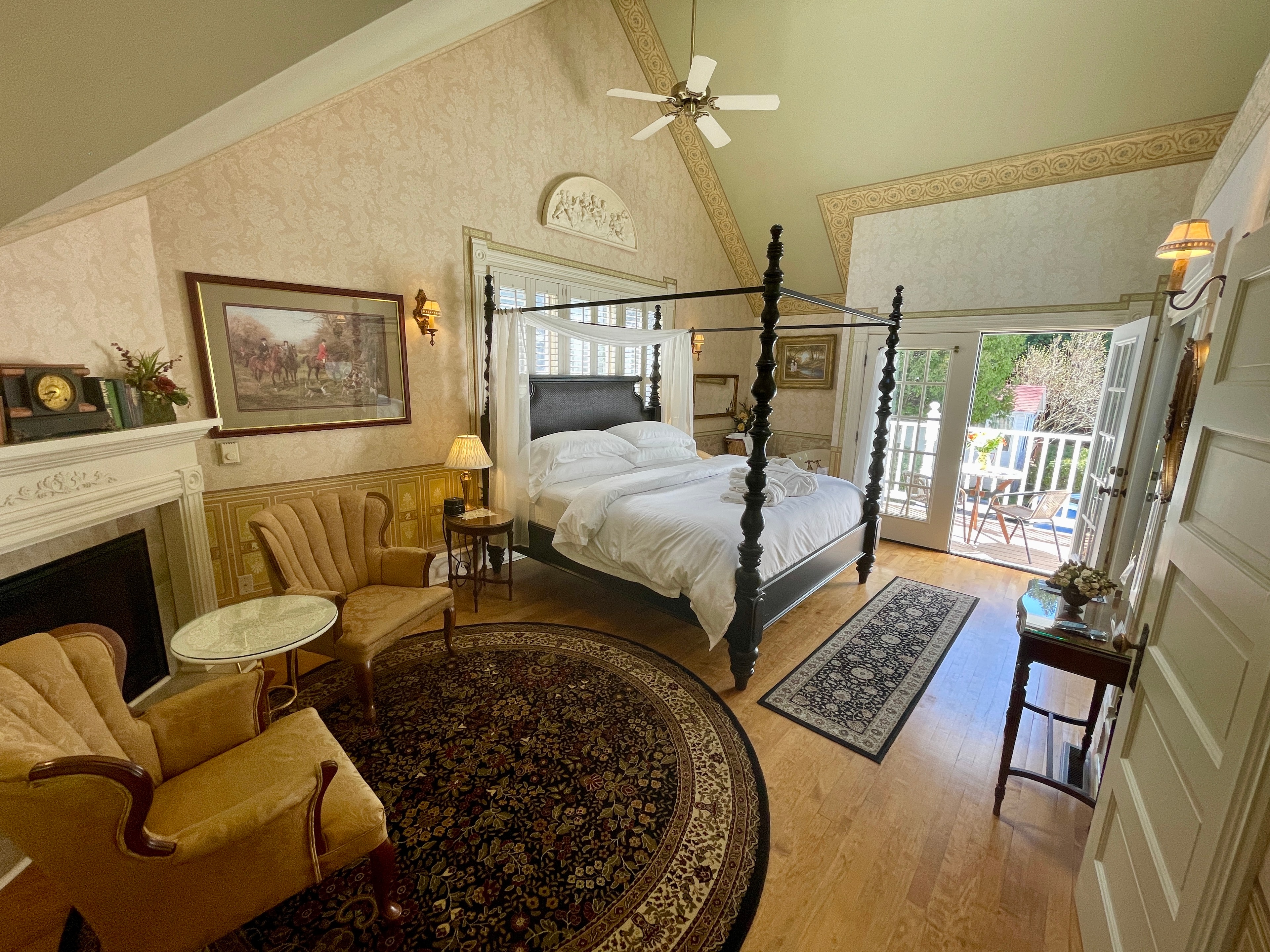 A grand bedroom featuring a dark wood four-poster bed, a fireplace, and a sitting area, with French doors leading to a balcony.
