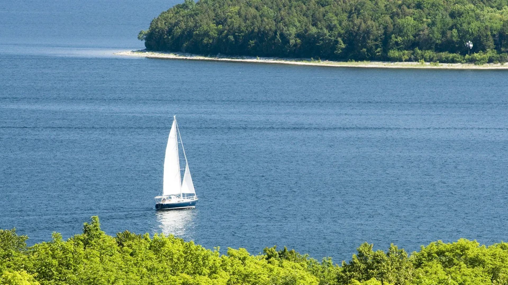 A sailboat glides across a calm blue lake near a green shoreline.