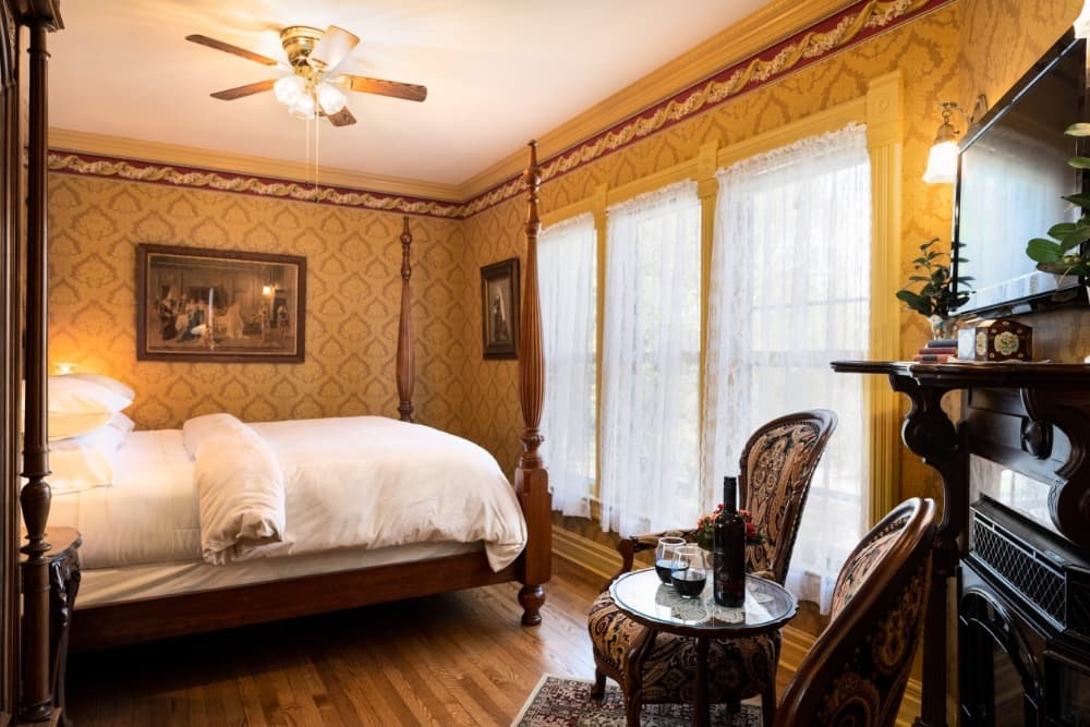 A cozy bedroom featuring a four-poster bed, antique decor, large windows with sheer curtains, and a small sitting area with wine glasses.