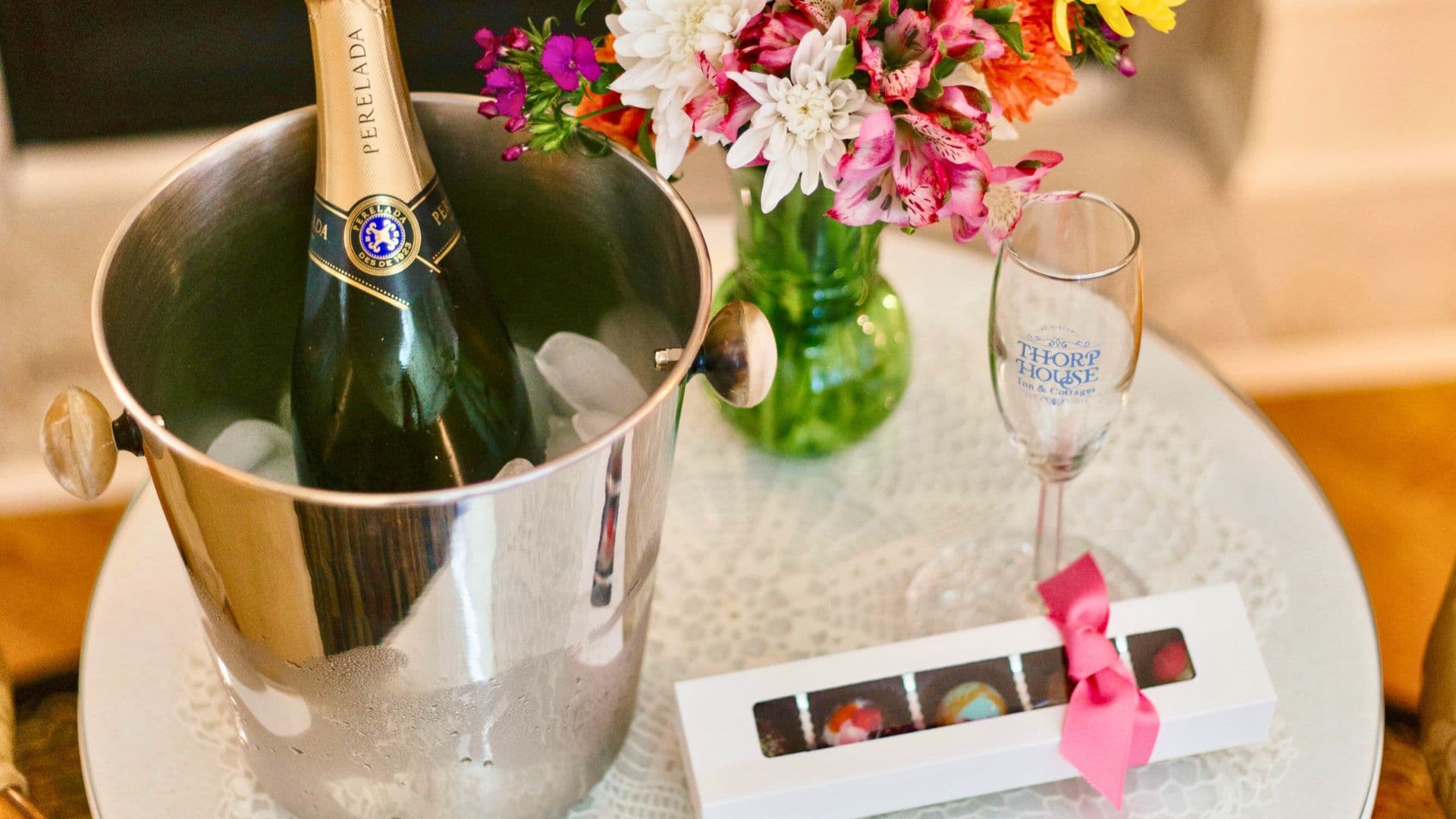 A bottle of champagne in an ice bucket is accompanied by a floral arrangement, a champagne flute, and a box of chocolates.