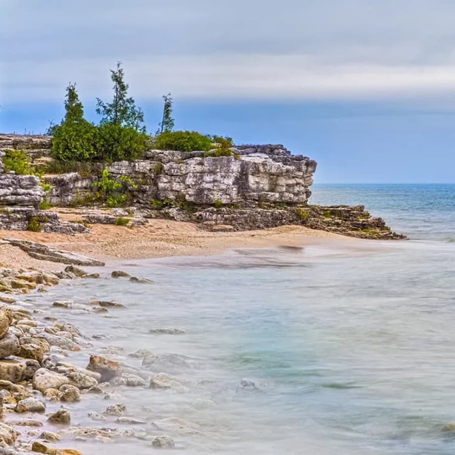 A rocky shoreline with sparse vegetation meets a calm, blue ocean under a cloudy sky.