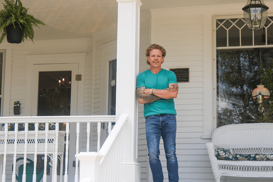 A man stands on the porch of a house, smiling and posing with his arms crossed.