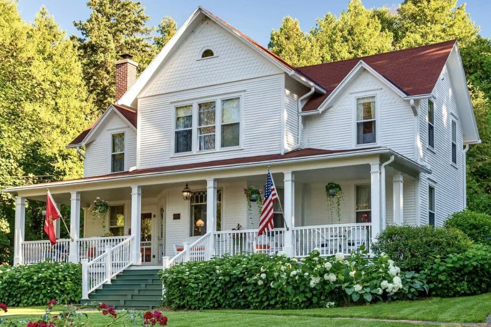 A charming white house with a red roof, surrounded by lush greenery and flowers, featuring a front porch with American and military flags.