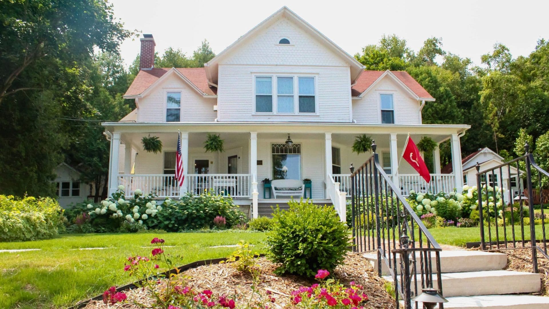 A charming white Victorian house with a porch, surrounded by lush greenery and flowers.