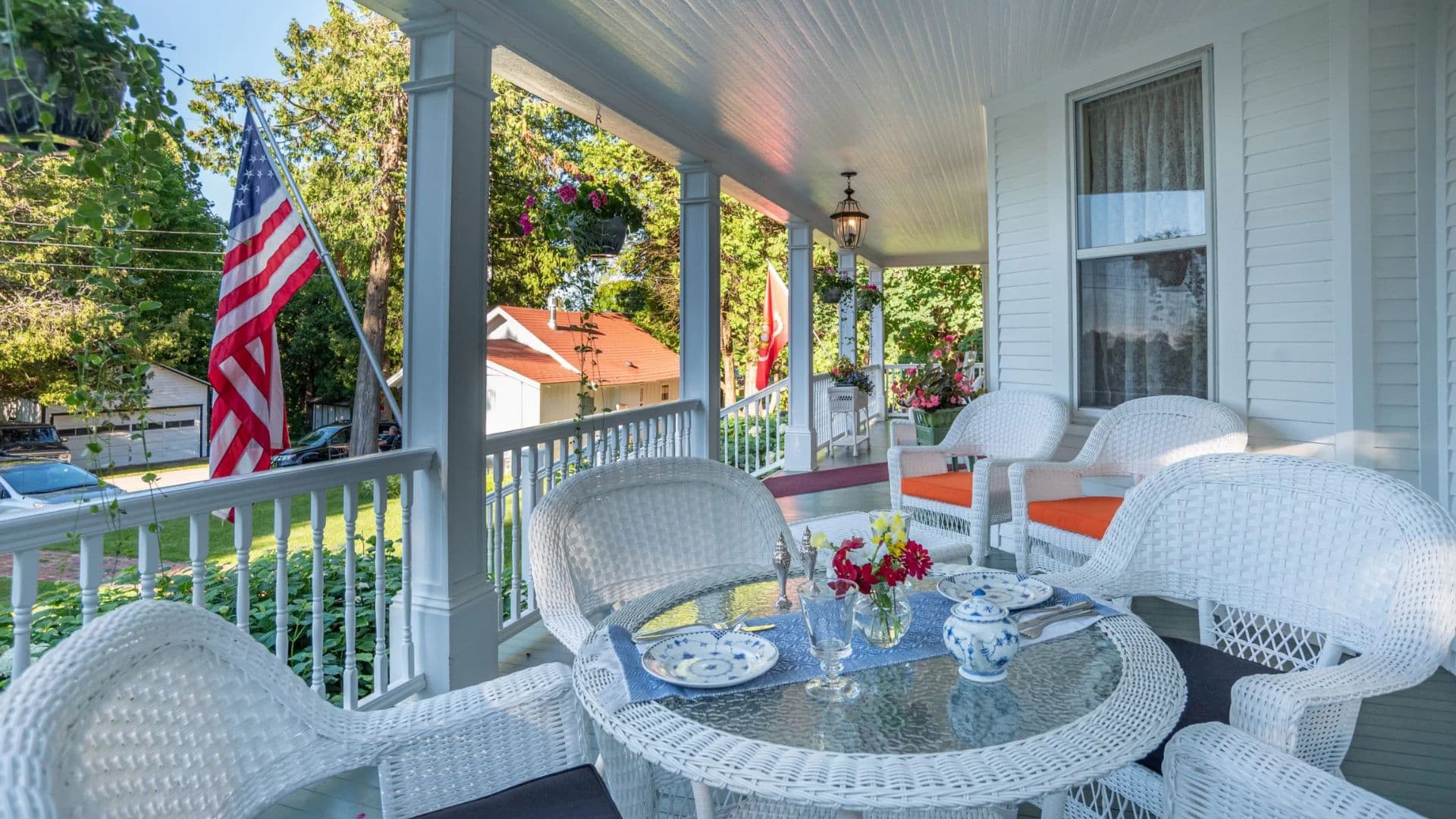 A quaint porch adorned with white wicker furniture, a glass table set for tea, and American flags in the background.